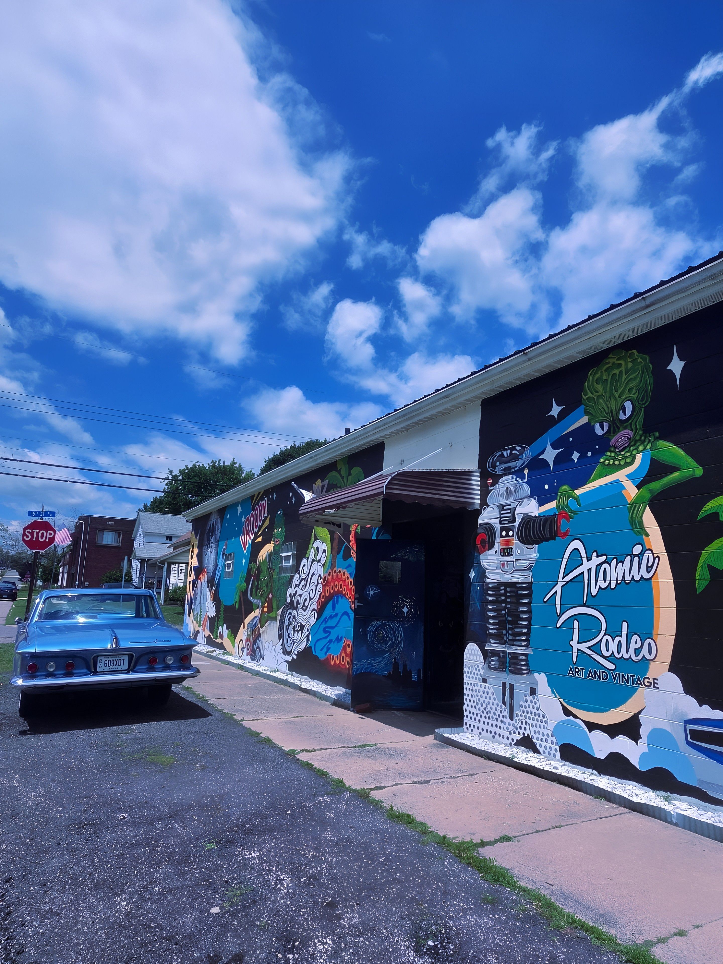 Bright blue sky with white clouds above a mural-covered storefront. A light-blue vintage car is parked on the street. The wall shows colorful sci-fi figures and the title "Atomic Rodeo".