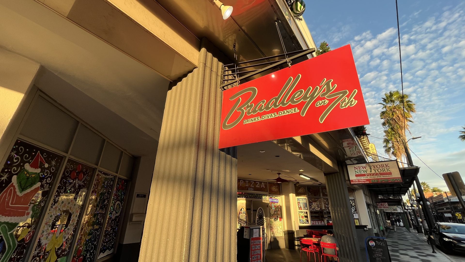 Red sign for Bradley's on 7th with yellow text, beige building columns, painted holiday windows, a few red chairs, palm trees, and a blue sky with clouds on a sunny day.