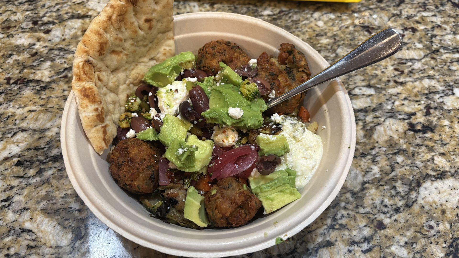 Bowl with falafel, avocado chunks, black olives, pickled onions, feta cheese, hummus, grilled veggies, and a piece of pita bread on a granite countertop with a spoon.