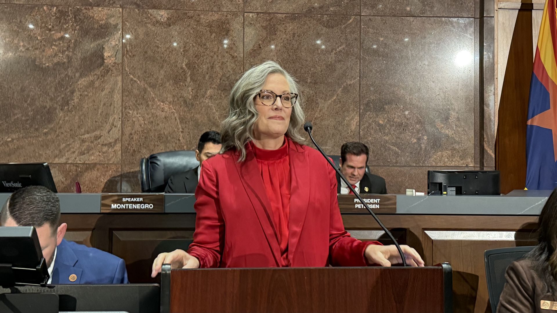 A woman in glasses wearing a red jacket speaking at a wooden podium with a microphone, with people seated behind and to the sides of her her, and an Arizona flag to the right.