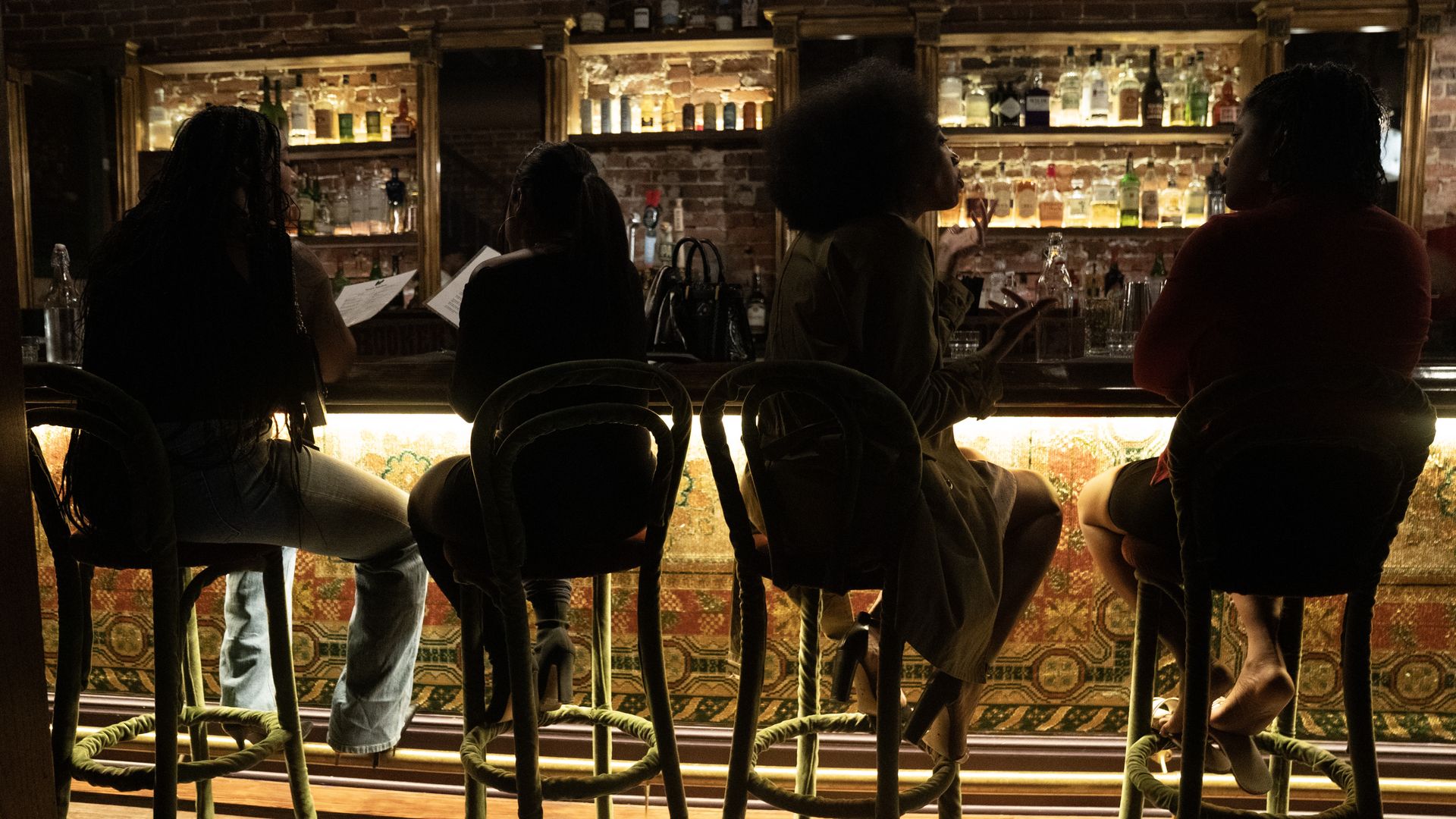Dim bar scene with four patrons seated along a wooden counter, facing bottles on backlit shelves. Silhouettes against exposed brick; green-legged stools with rope footrests and warm lighting.