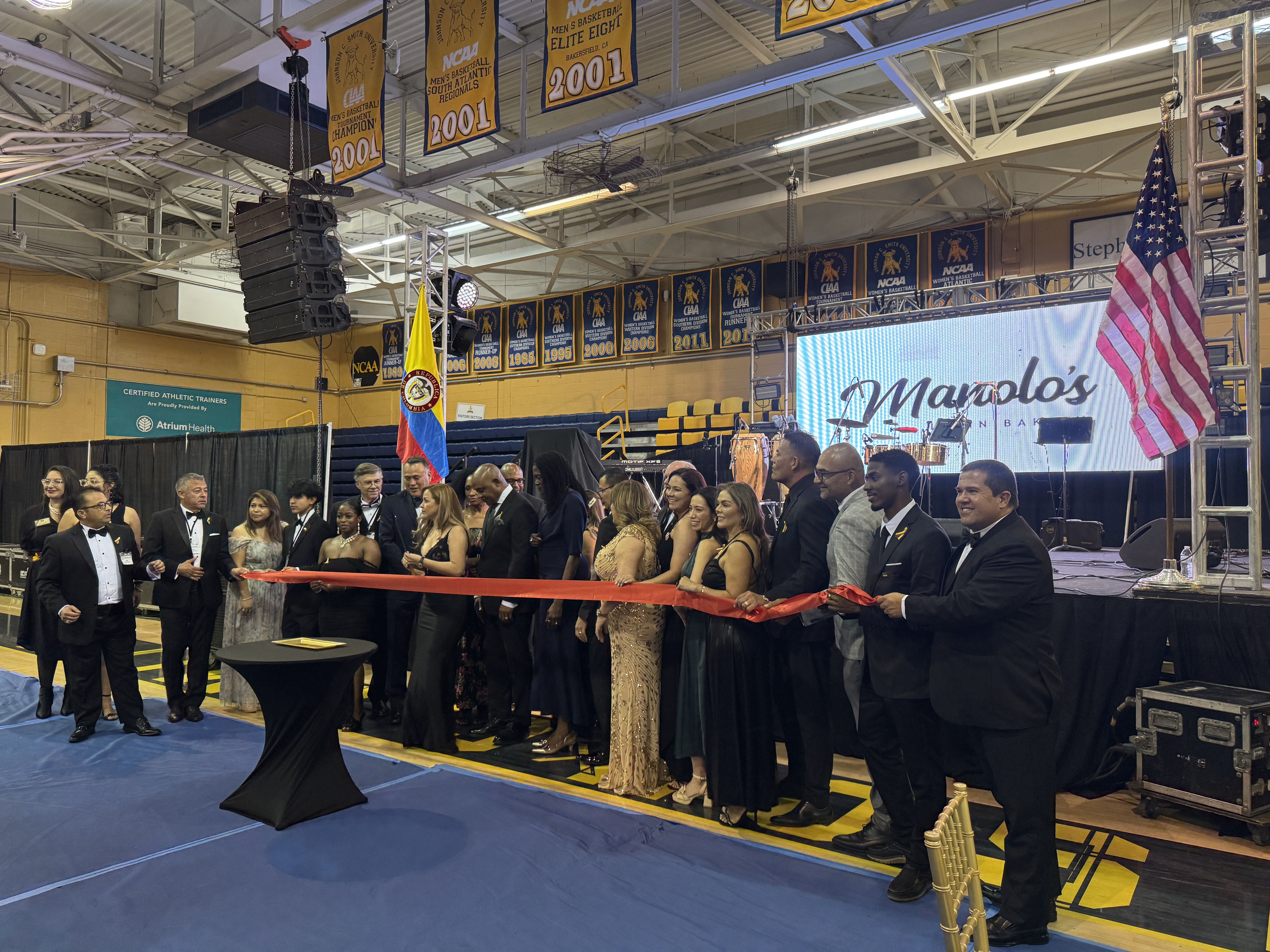 A group of people dressed in formal attire participates in a ribbon-cutting ceremony inside a gymnasium. The backdrop includes banners and flags, and a stage with an LED screen displaying 'Marolo's' is visible. The setting suggests a celebratory event or opening."