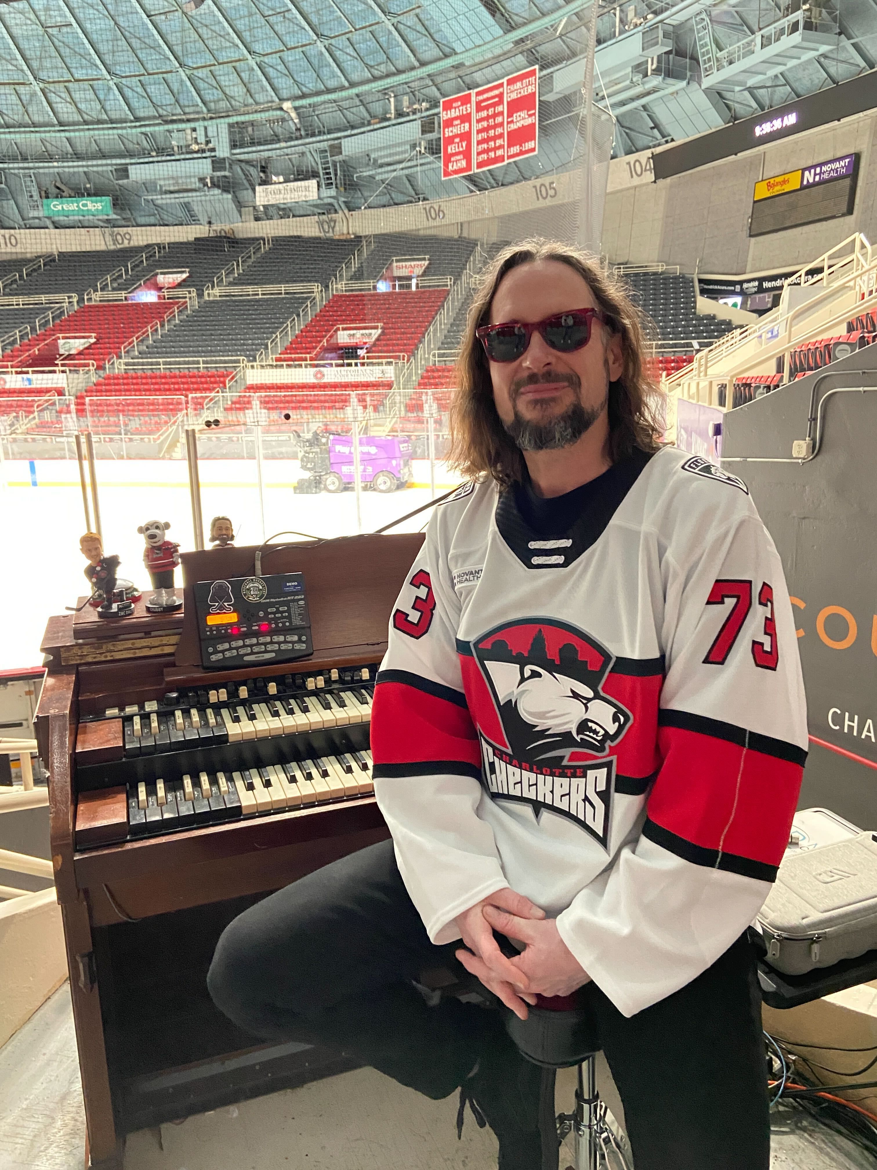 Man with shoulder-length hair and beard wearing sunglasses and a white Charlotte Checkers hockey jersey with number 73 sits on a stool next to an organ in an empty indoor ice rink.