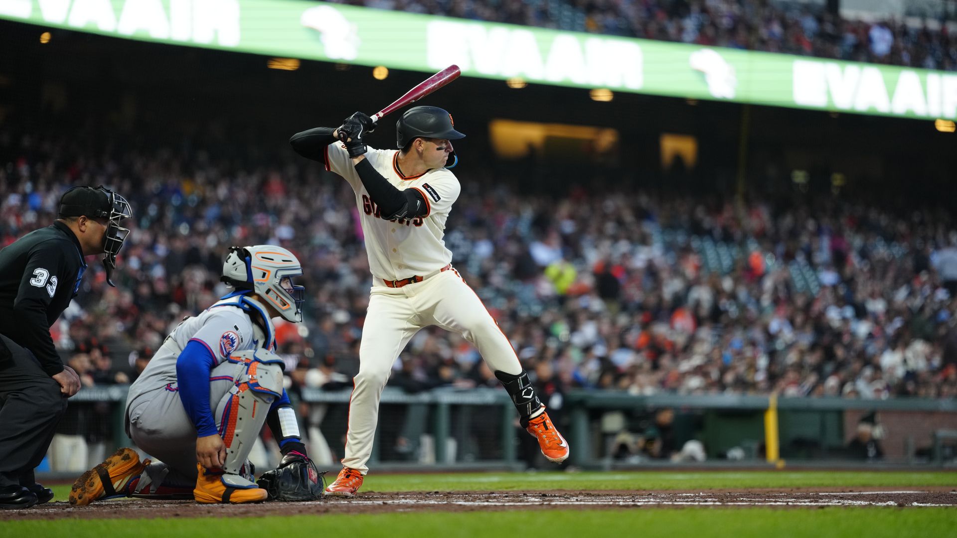 Baseball game: a batter in a white Giants uniform swings a red bat as the catcher and umpire watch; a crowded stadium glows with green signage in the background.