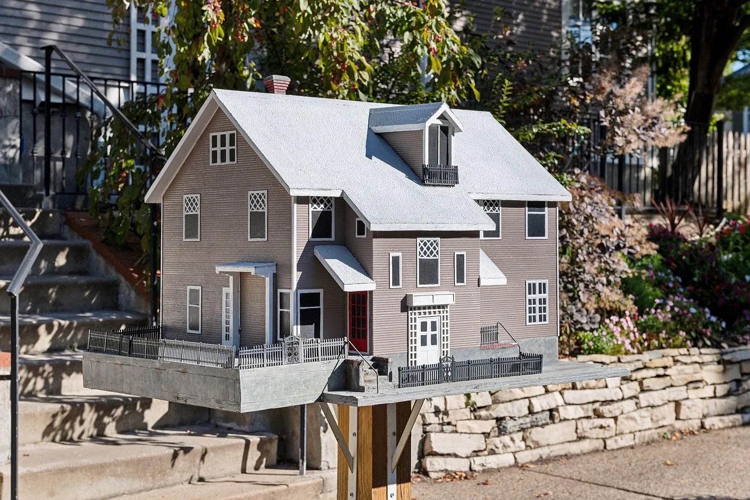 Detailed miniature model of a two-story beige house with white trim and a red door, set on a wooden post, with black fences and realistic windows, in an outdoor garden area.