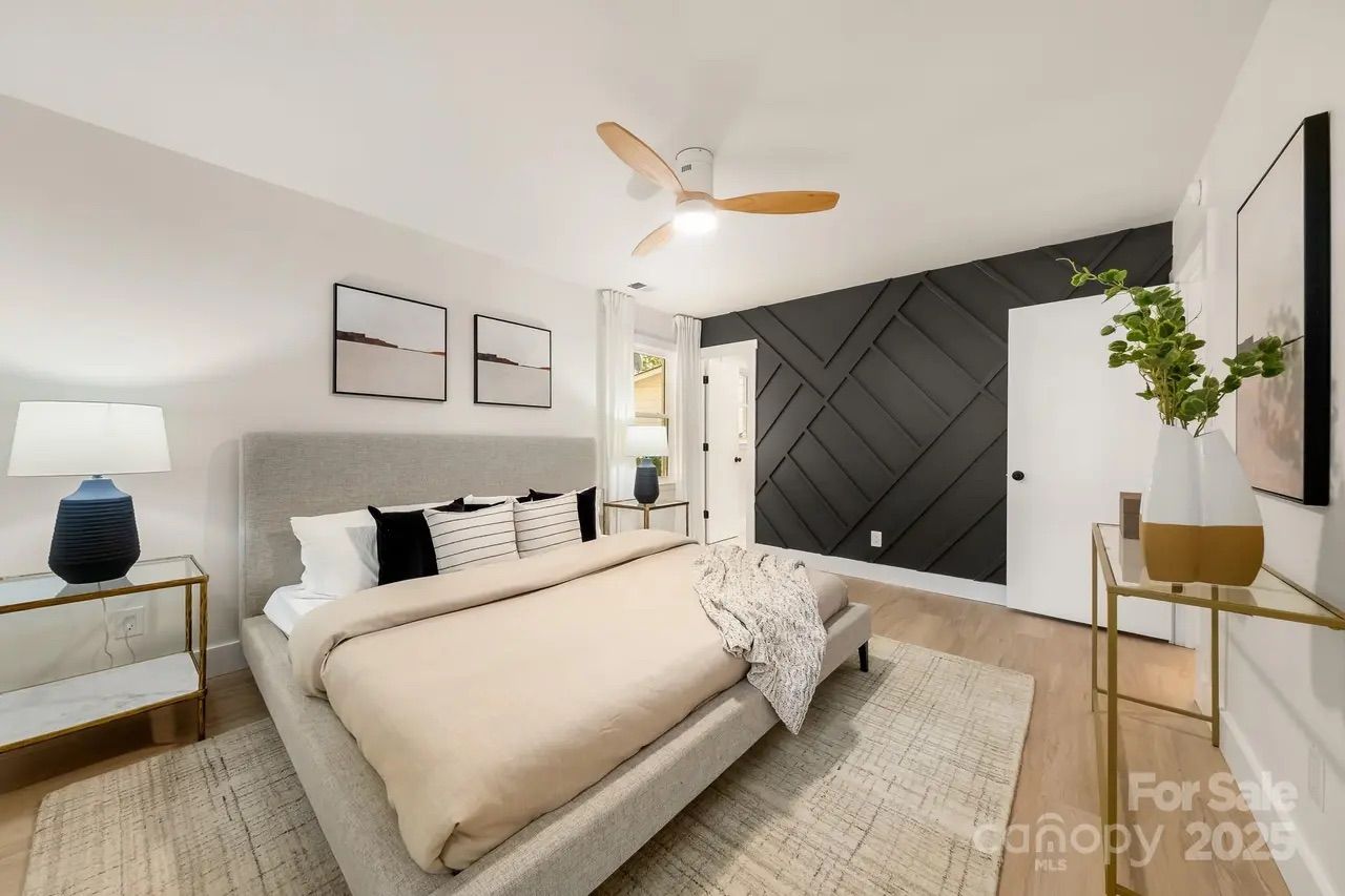Modern bedroom with beige bedding, gray upholstered bed, two nightstands with blue lamps, a black accent wall with diagonal paneling, a ceiling fan, light wood flooring, and a simple vase with green leaves.