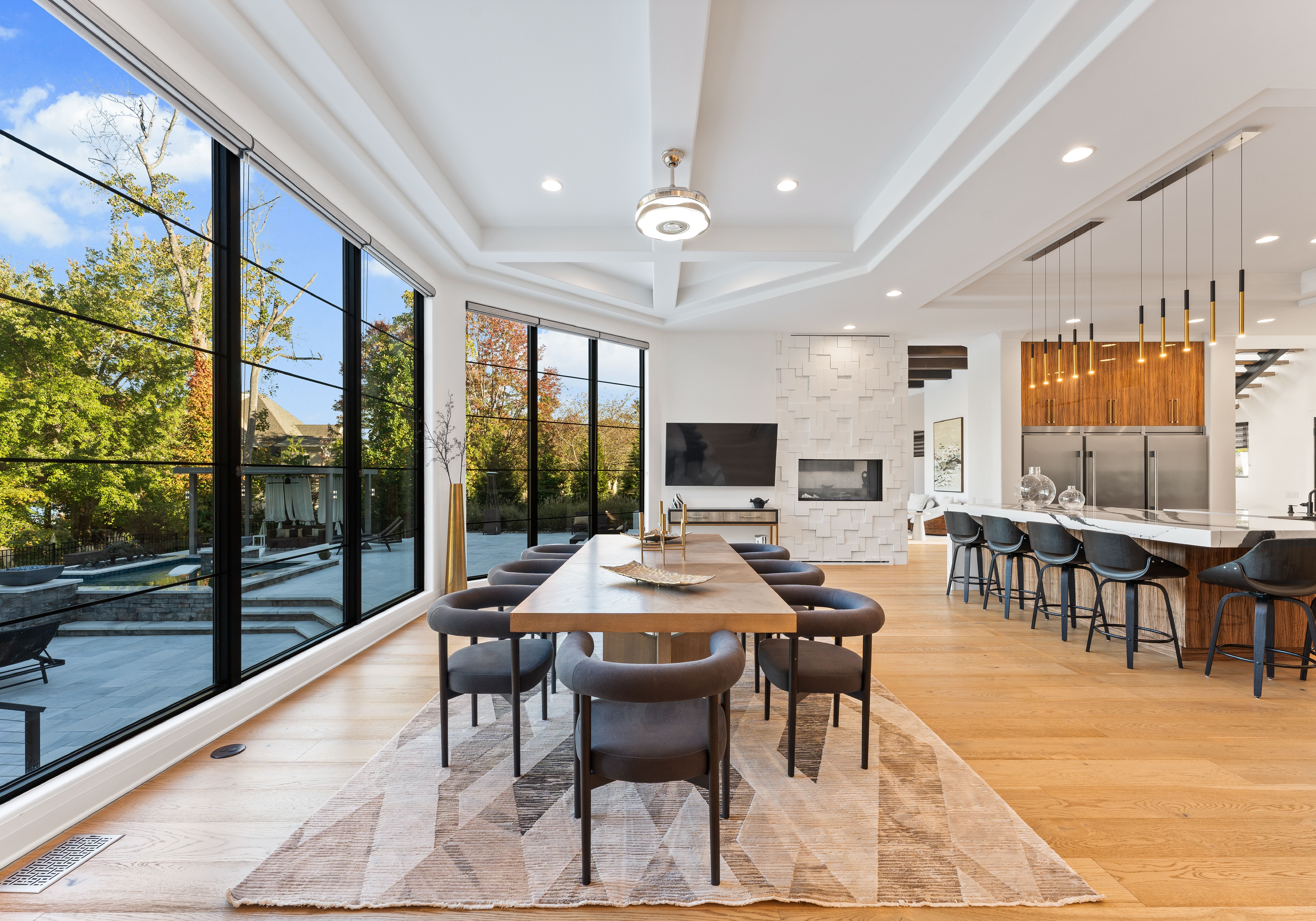 Modern dining room with long wooden table, eight black chairs, large windows showing trees and blue sky, geometric rug, white coffered ceiling, and kitchen area with bar stools and pendant lights.