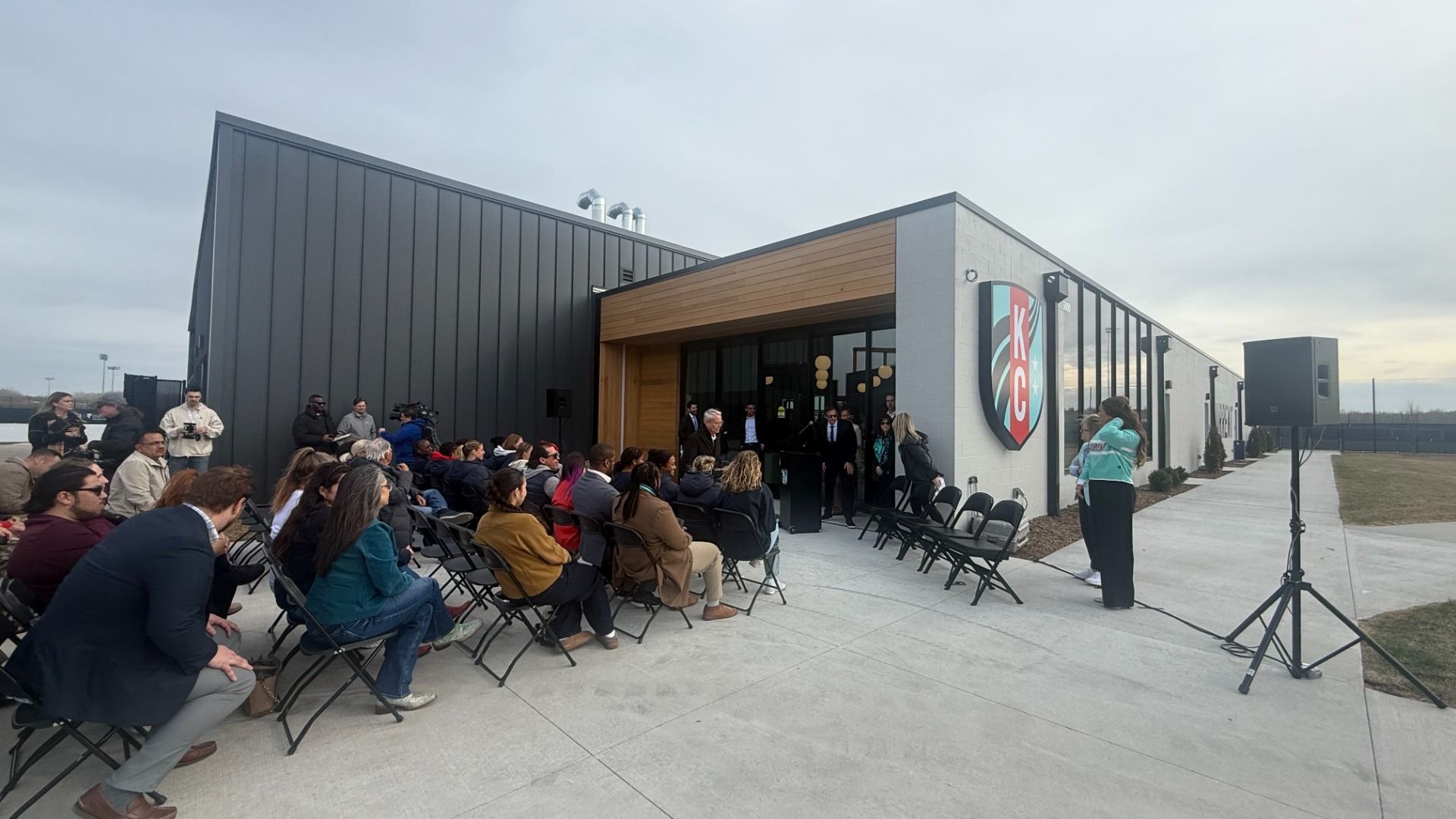 Outdoor event with people seated on black folding chairs in front of a modern building with a large KC shield logo, a speaker on a stand, and cloudy sky overhead.