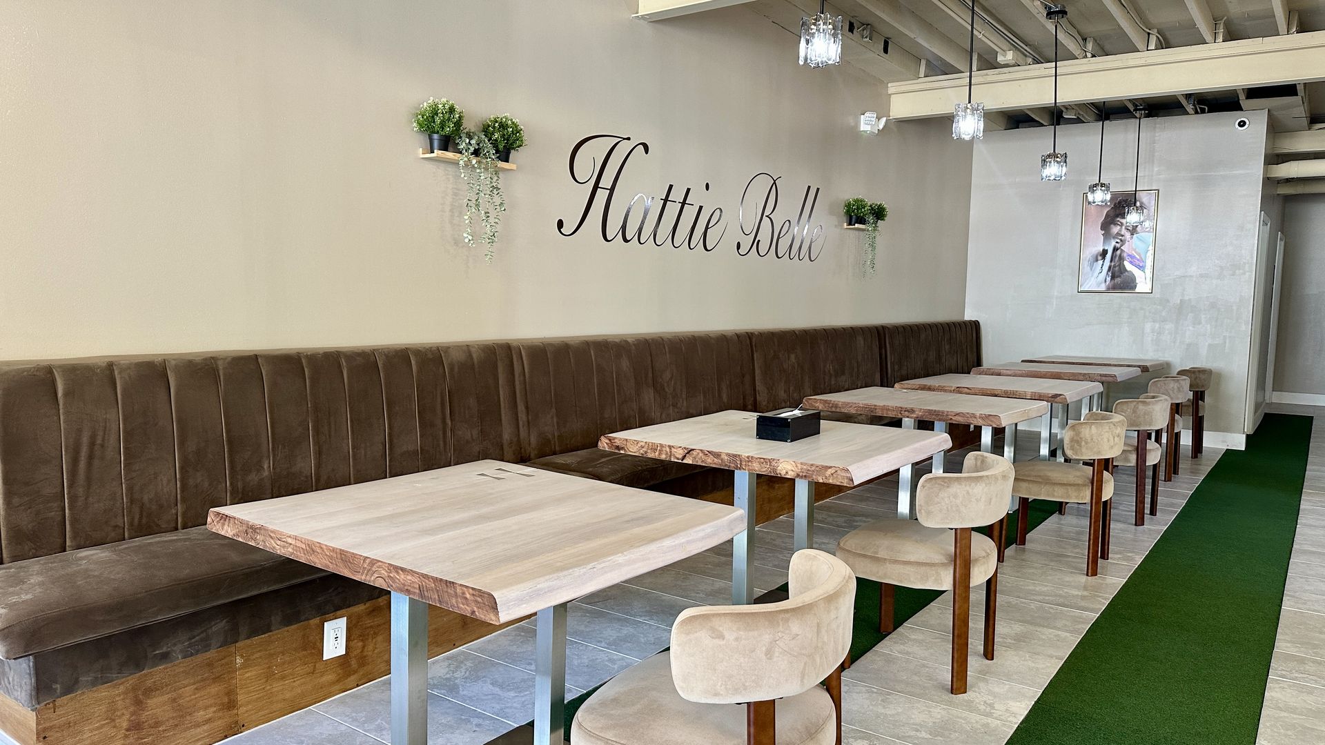 Cafe interior with a long brown bench along a wall that reads "Hattie Belle" in cursive. Plants on shelves, wooden tables, beige chairs, and hanging pendant lamps; framed poster on the wall.