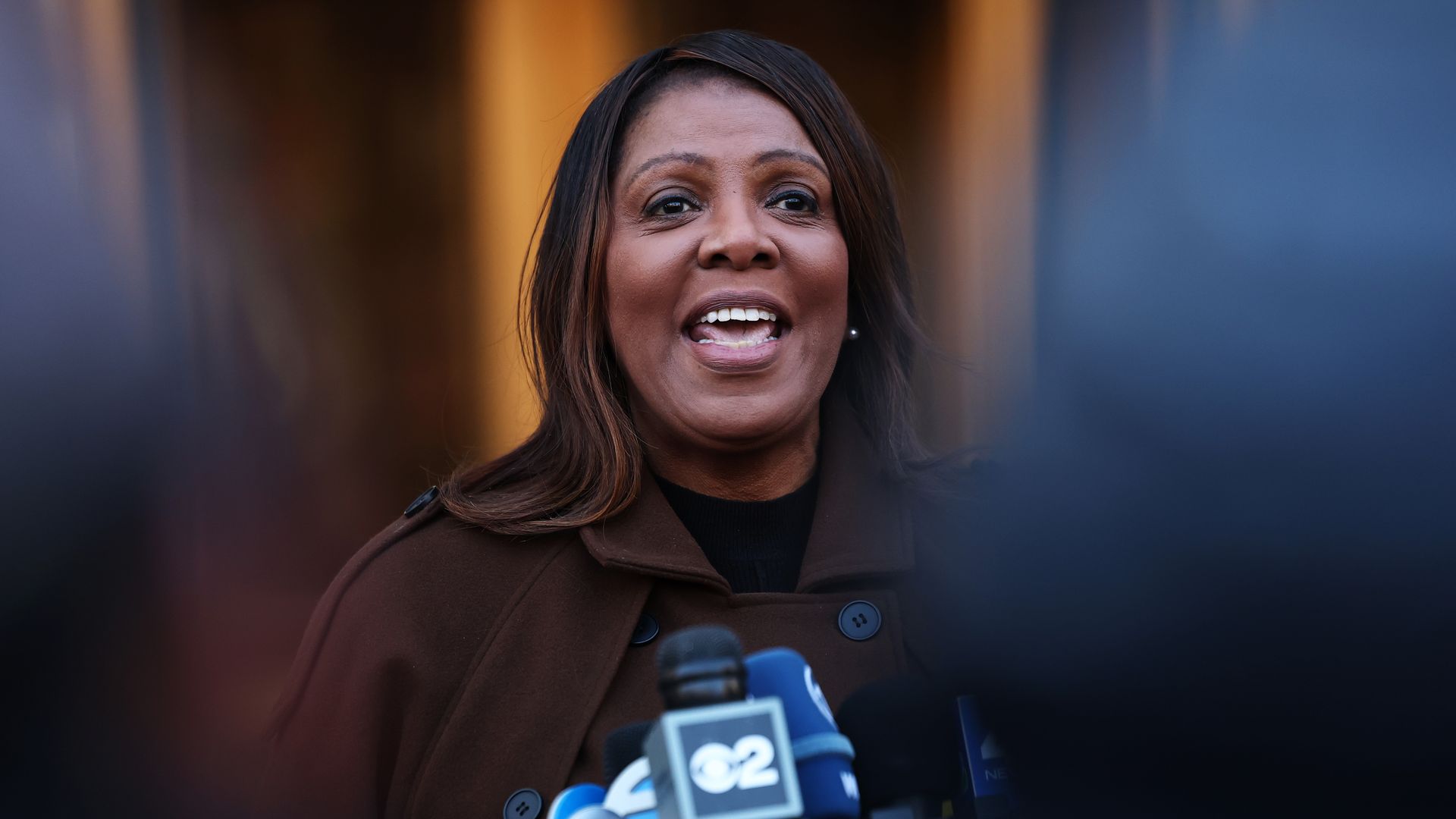 Black woman wearing a brown coat speaks at a press conference with multiple microphones from news channels like abc and cbs in front of her.