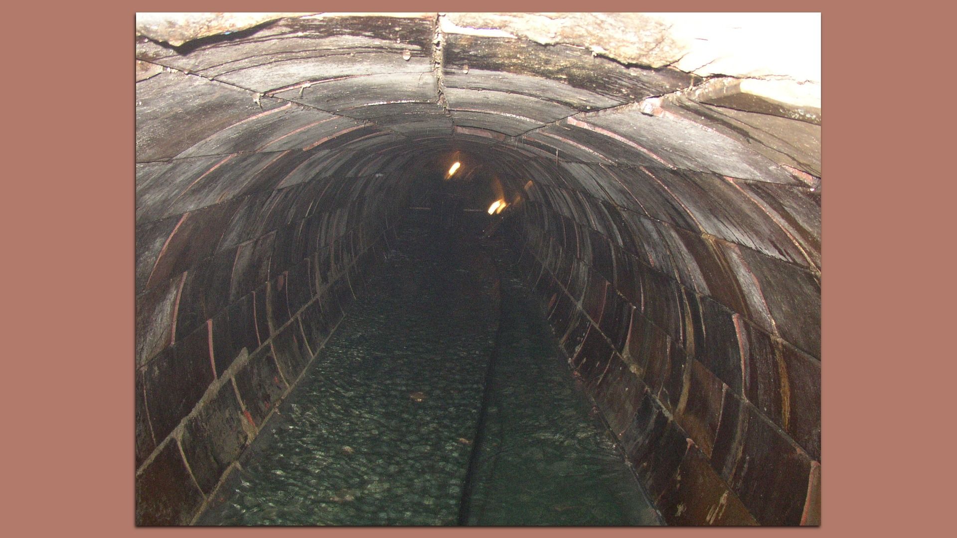 Dark cylindrical brick tunnel with arched, damp walls and a water-filled floor; a few orange lights glow faintly far down the tunnel.