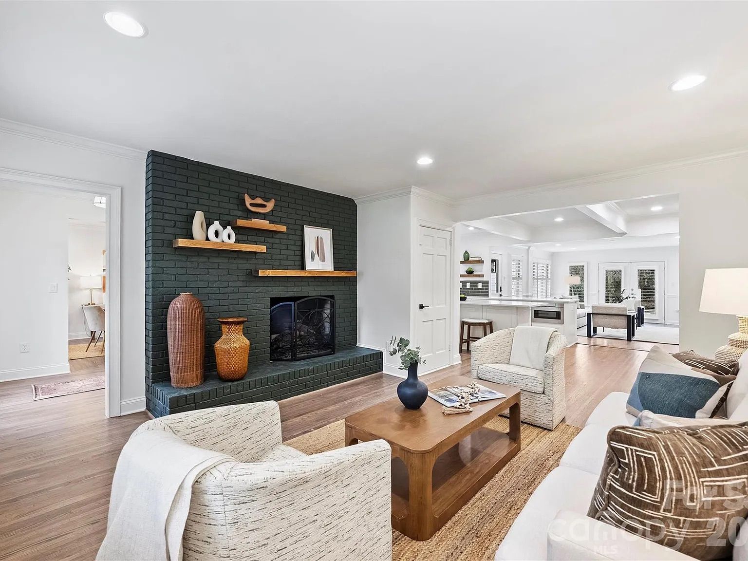 Bright living room with dark green brick fireplace, wooden floating shelves, wicker vases, beige armchairs, wooden coffee table, white sofa with patterned cushions, and light wood floors.