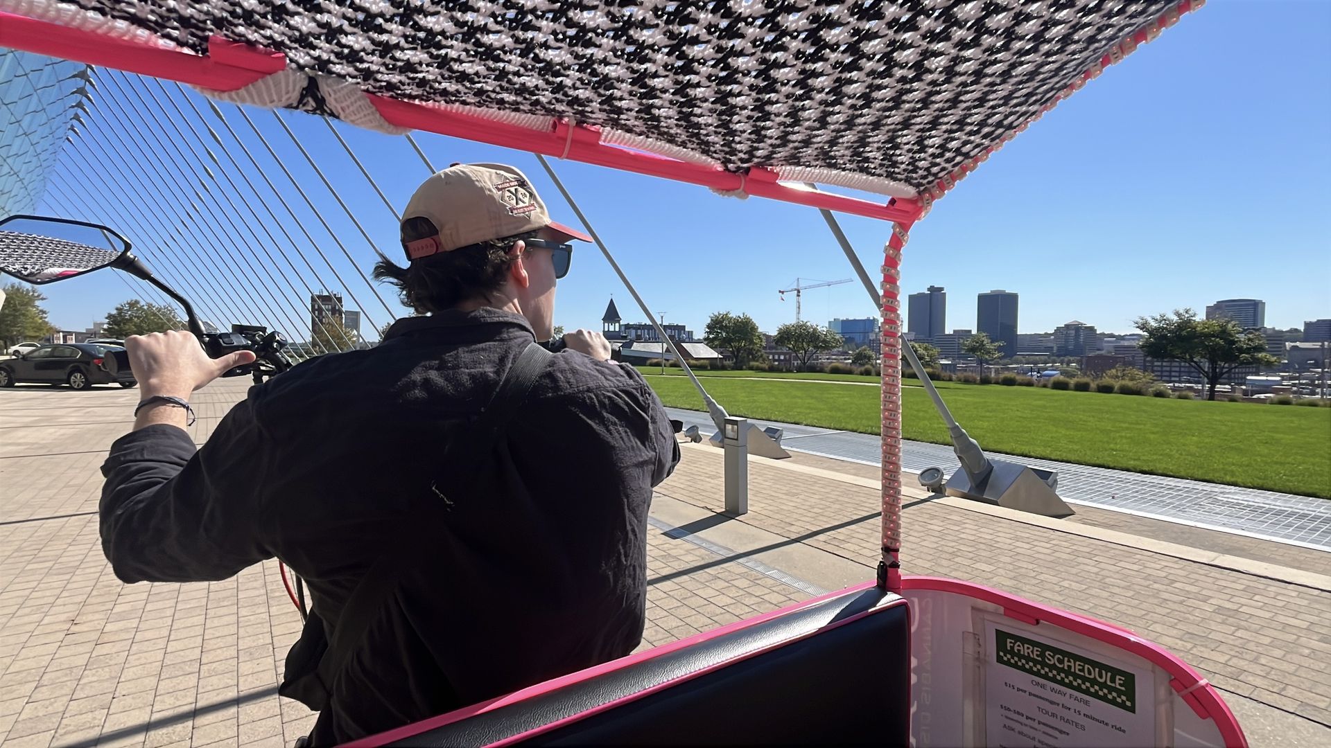 Mark Manning, director of operations with E-Z Pedicabs, wearing a beige cap and black shirt drives a pink pedicab on a sunny day near the Kauffman Center and looks out over the Crossroads.