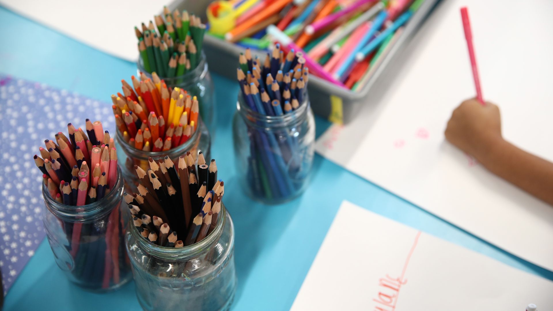 Piles of colored pencils are shown on a desk next to a child coloring