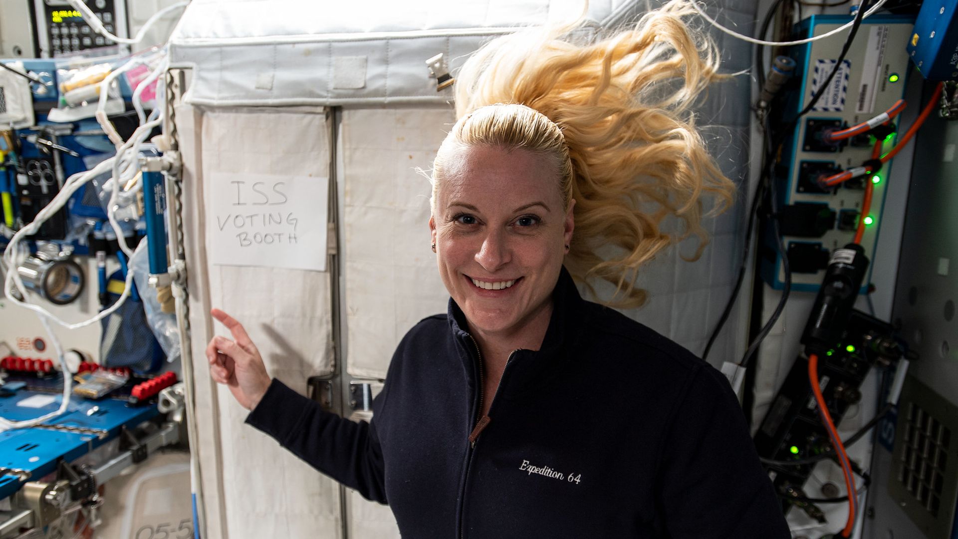 Astronaut Kate Rubins floating in front of a sign that says "ISS voting booth" 
