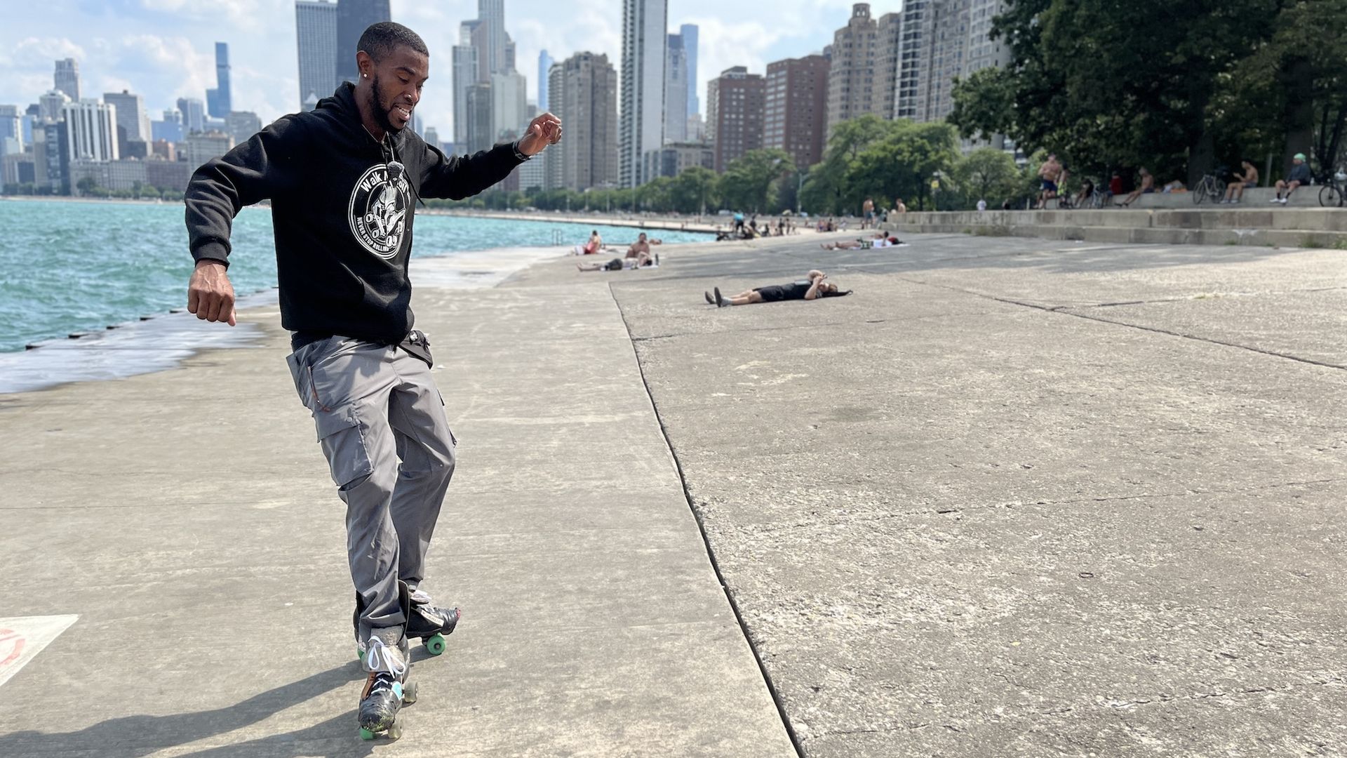 Brandon Mangrum in gray pants and a black hoodie rollerblades near a lakeside promenade with the Chicago skyline and people relaxing in the background on a sunny day.