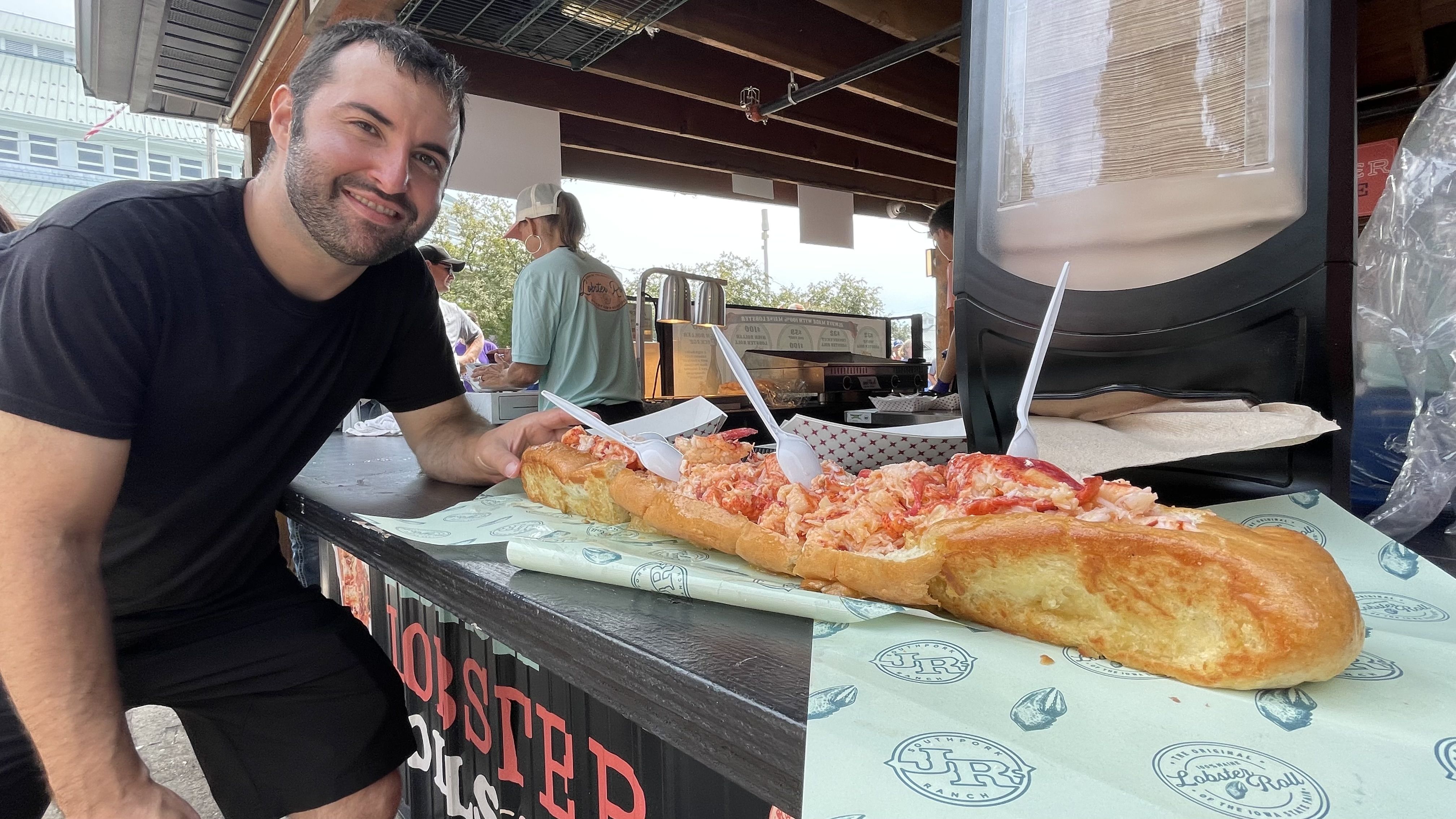 Smiling man in black shirt beside a very long lobster roll sandwich on a counter with branded paper, outdoors at a food stand with people in the background.