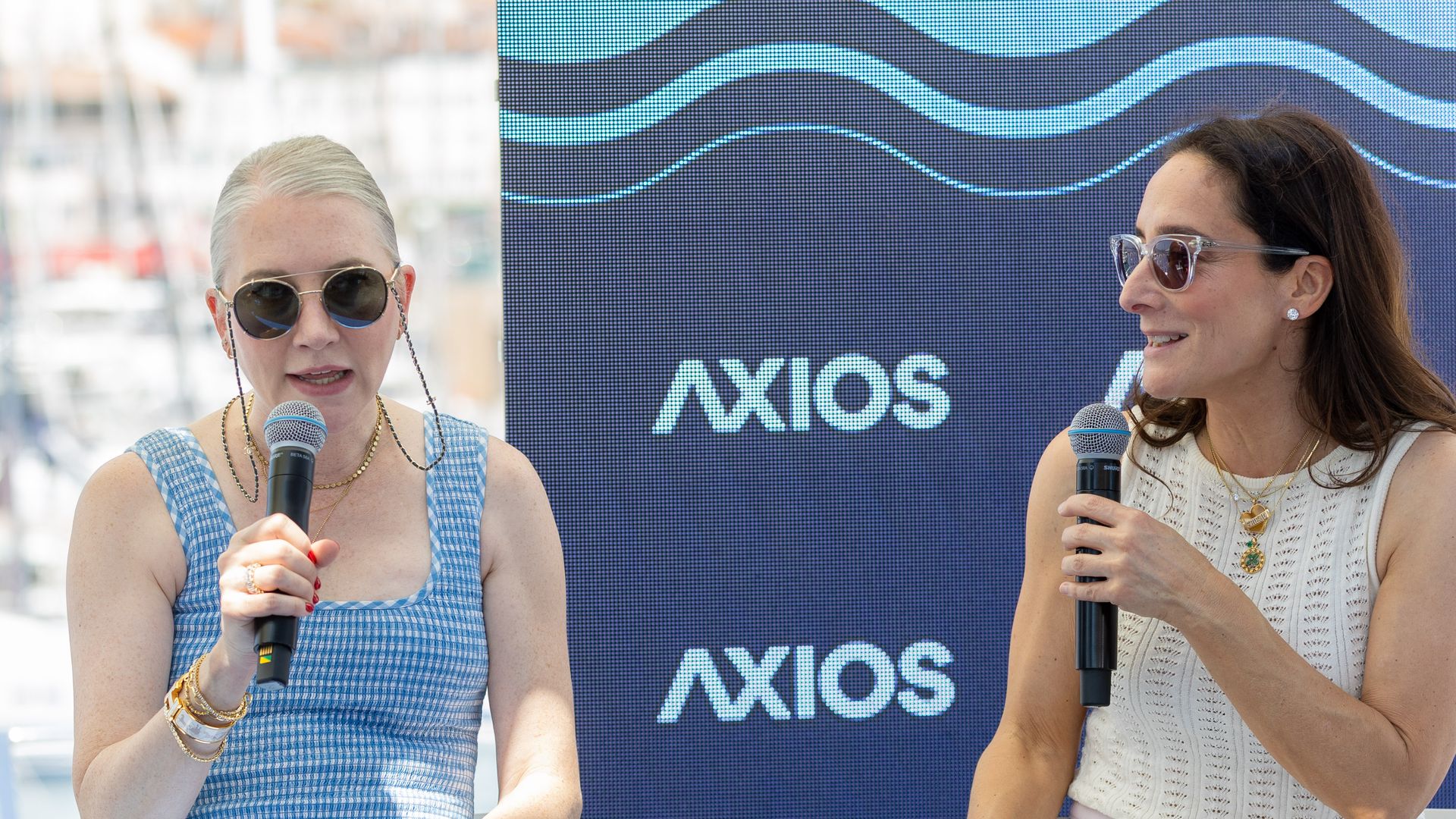 Two women in sunglasses hold microphones at an outdoor event, speaking in front of a blue digital backdrop showing waves and the word AXIOS. Left, silver hair in a blue check dress; right, white top.