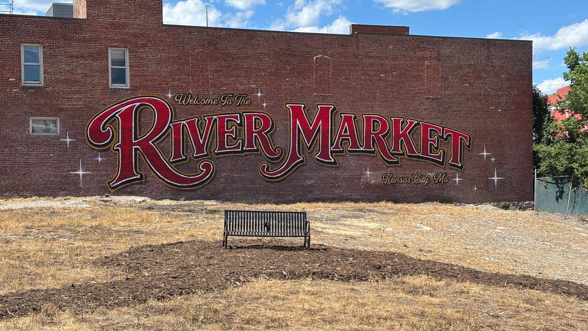 Brick wall mural reading Welcome To The River Market in large red letters with gold trim; Kansas City, MO; bench on dry grass, blue sky above.