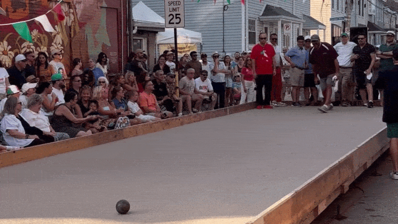 Crowd gathers along a makeshift wooden bocce court on a street; a dark bocce ball rests near the foreground as banners and a Speed Limit 25 sign appear in the background.