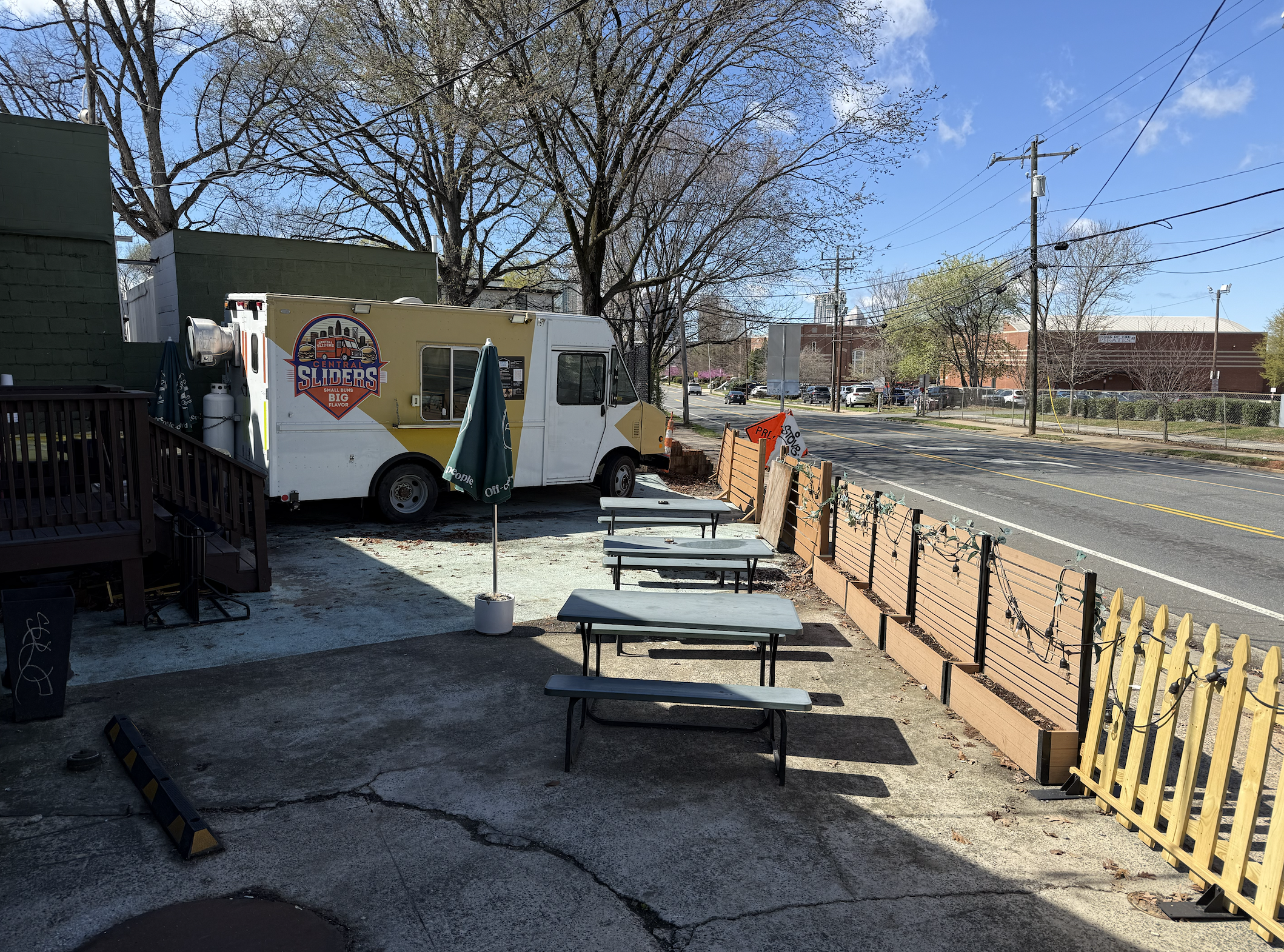 Food truck with a Central Sliders logo parked beside an outdoor patio. Three green picnic tables with umbrellas sit along a fence by the street, under a blue sky with bare trees and power lines.