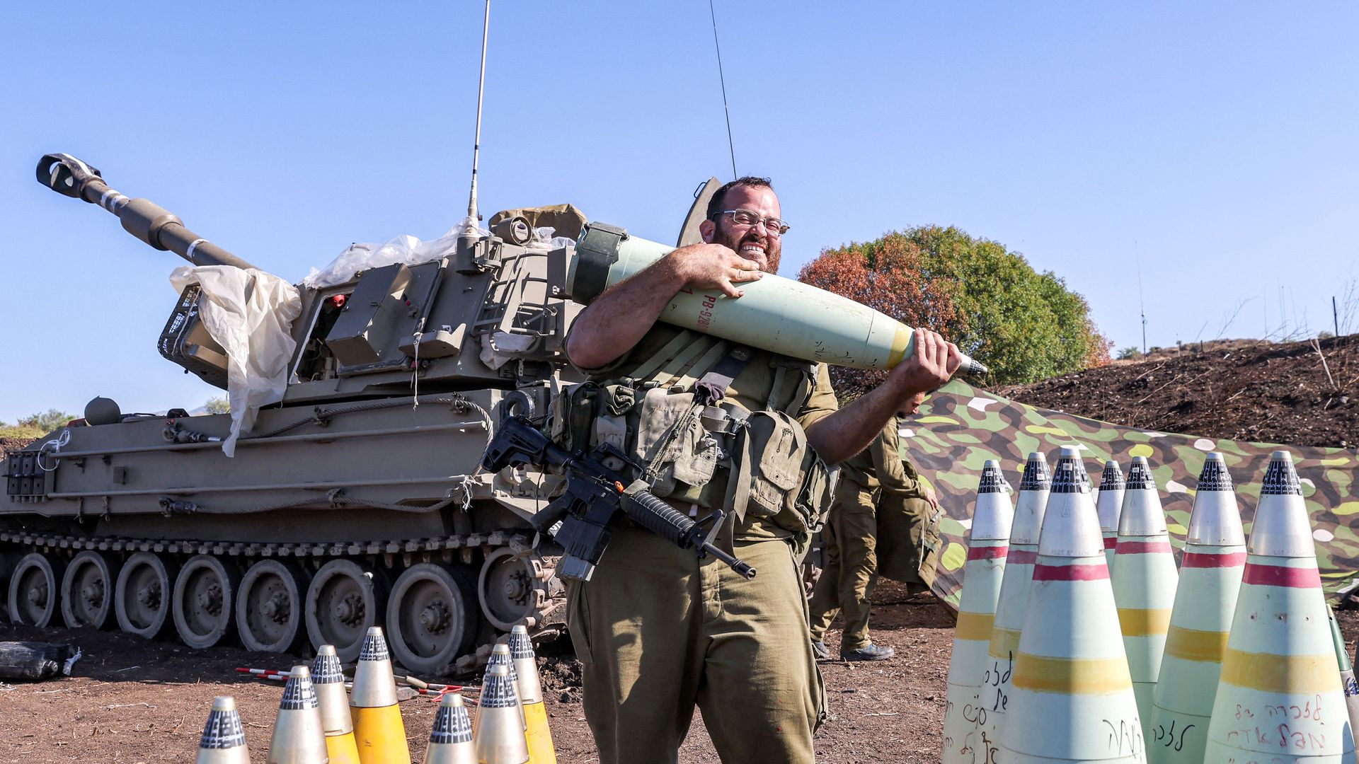 An Israeli soldier lifts a 155mm artillery shell with a self-propelled Howitzer in the background. 