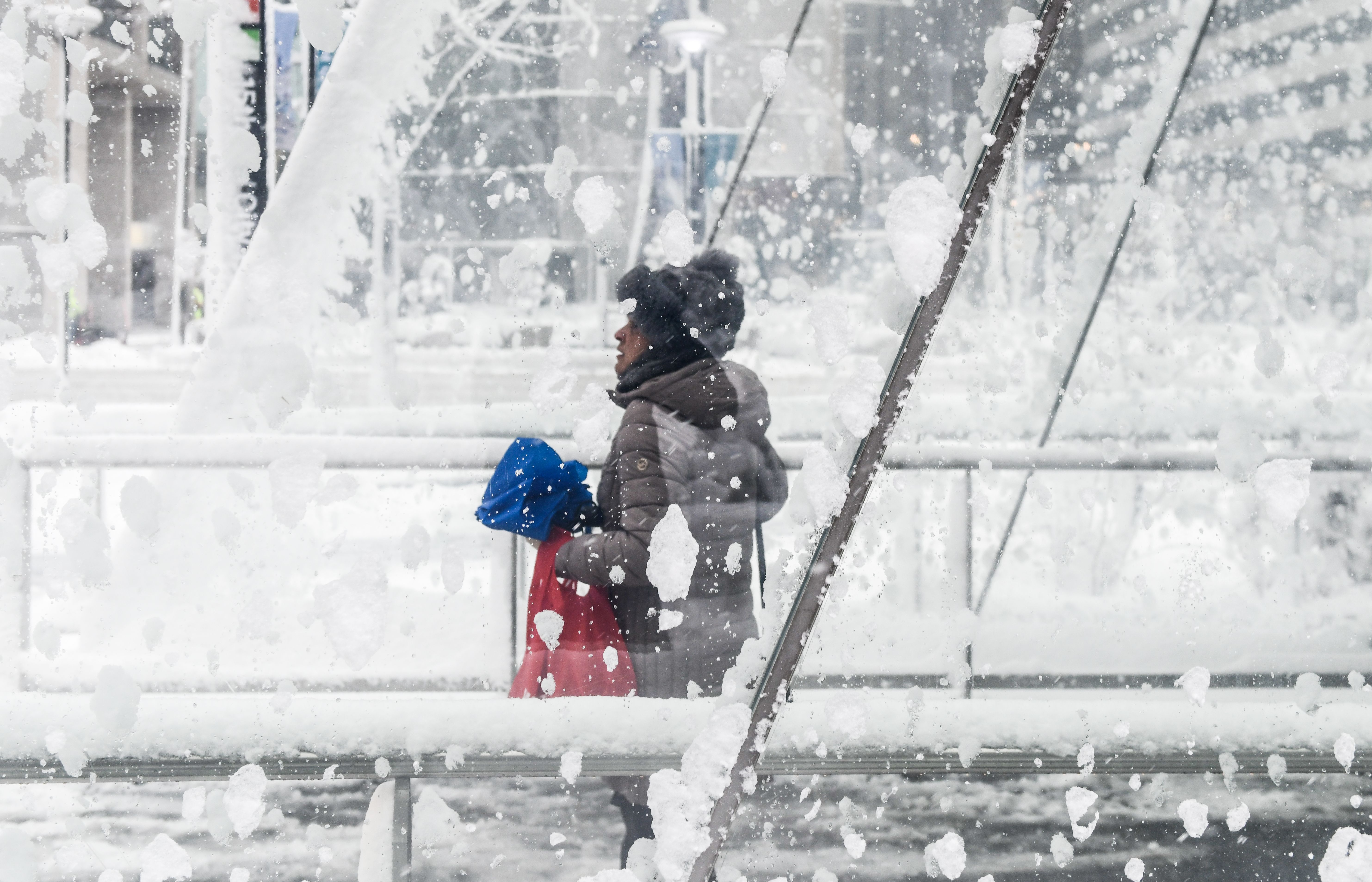 Person in a gray winter jacket and black fur hat holds blue umbrella and red bag, seen through a snow-covered glass window in a snowy urban setting.