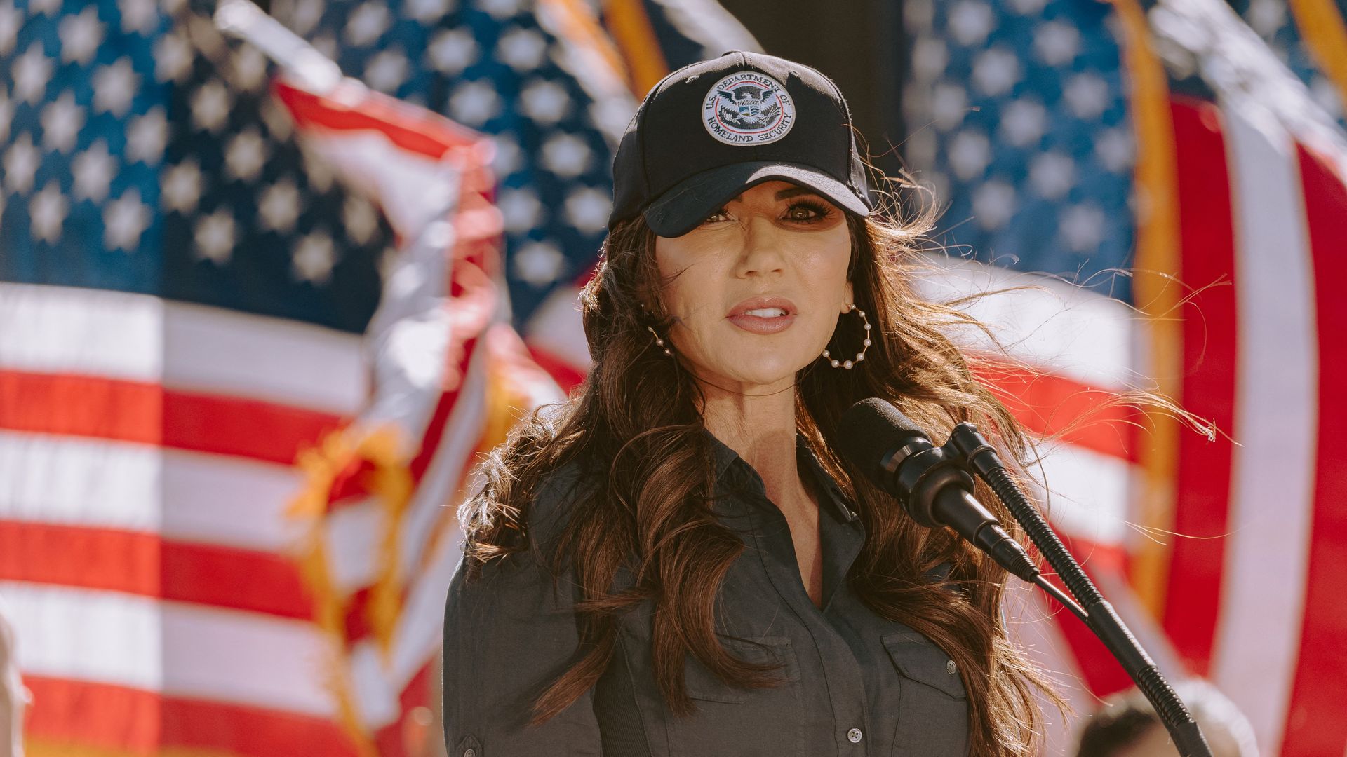 Woman with long brown hair speaking at a microphone, wearing a U.S. Department of Homeland Security black cap and dark button-up shirt, with American flags in the background.