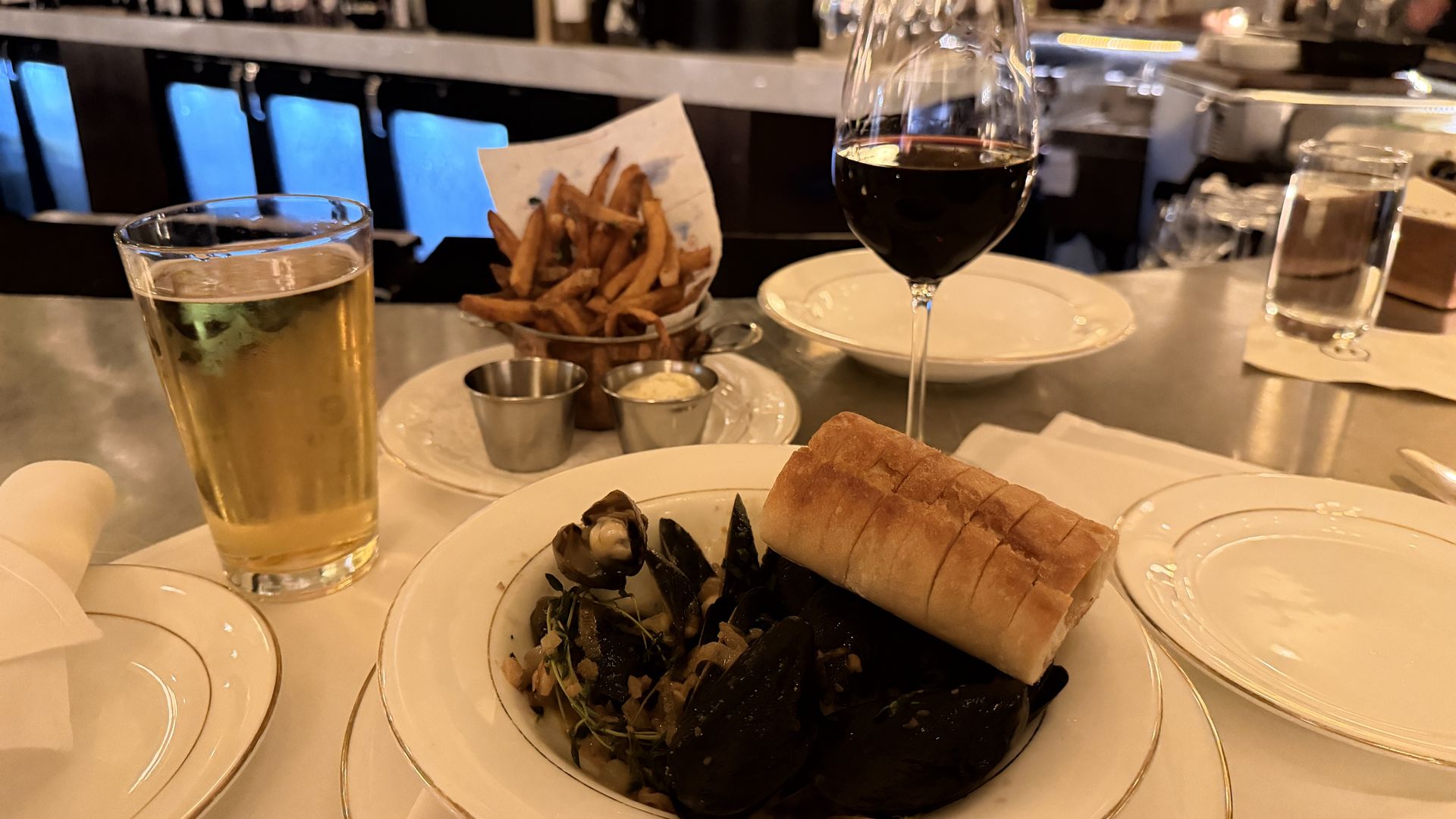 Mussels with a bread roll on top, served on a plate; pale beer glass to left and red wine glass to right, fries and sauces in a basket behind, bar with patrons in background.