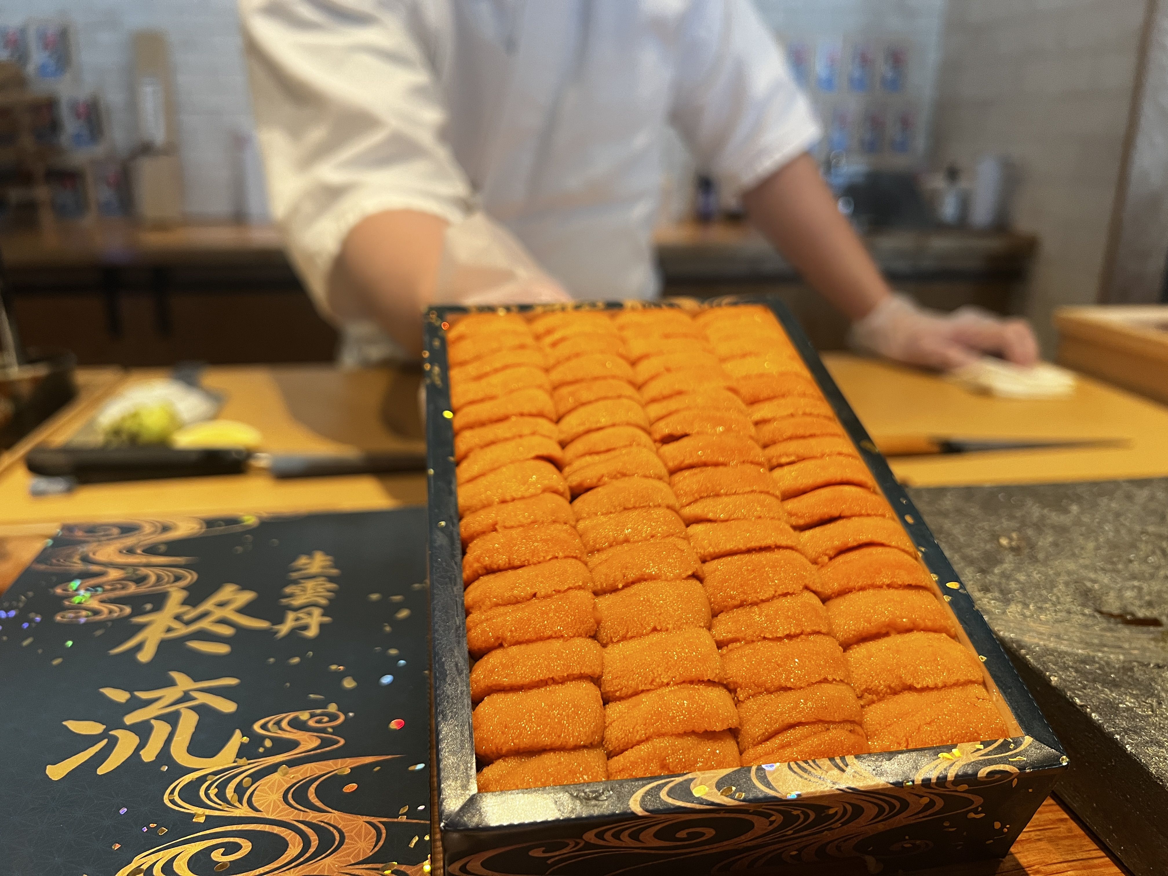 Chef in a white coat stands behind a kitchen counter, presenting a black box with gold scrollwork filled with neatly arranged bright orange round confections in rows.