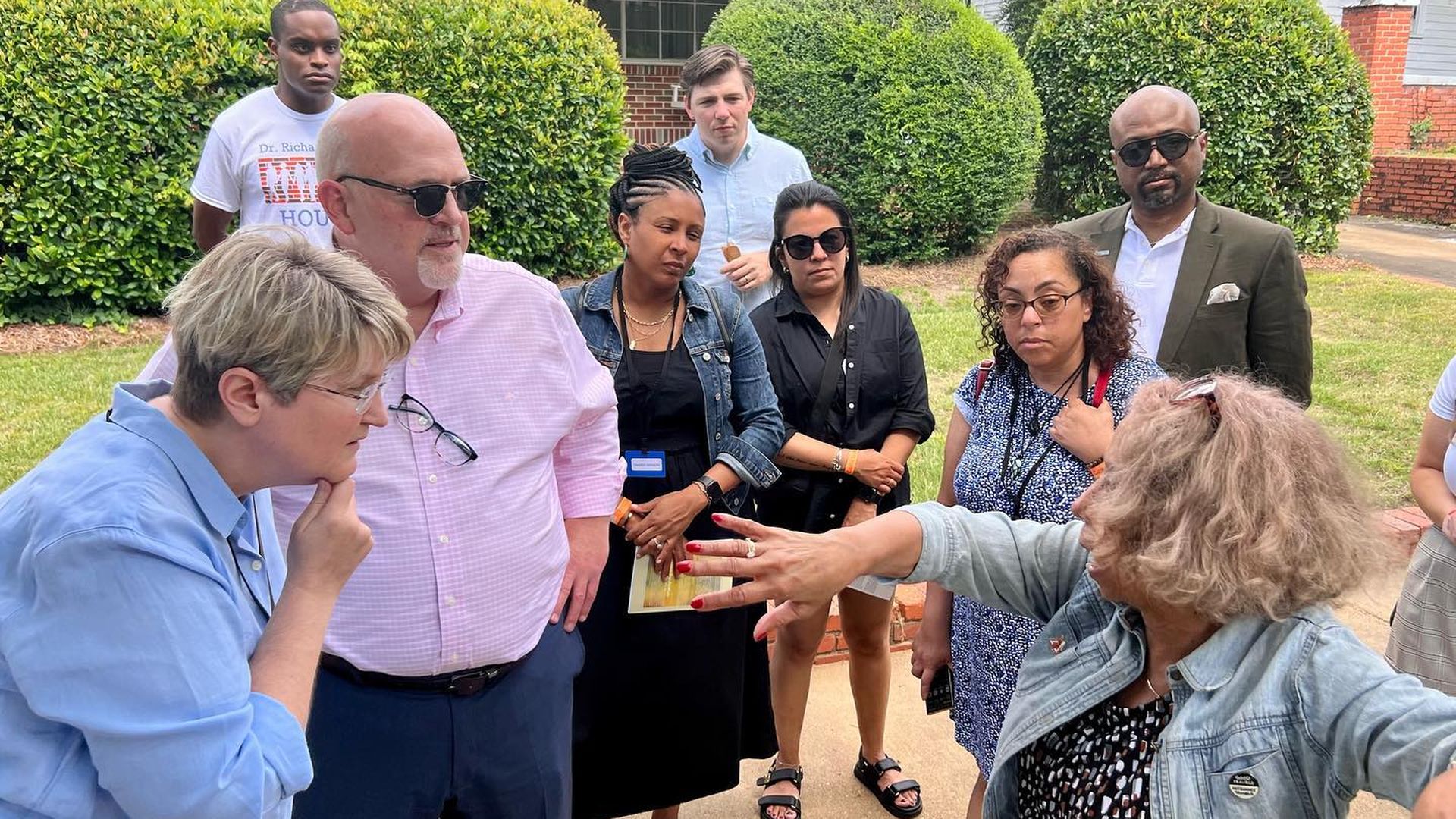 A group of people listens to a tour guide on the sidewalk in front of bushes. 