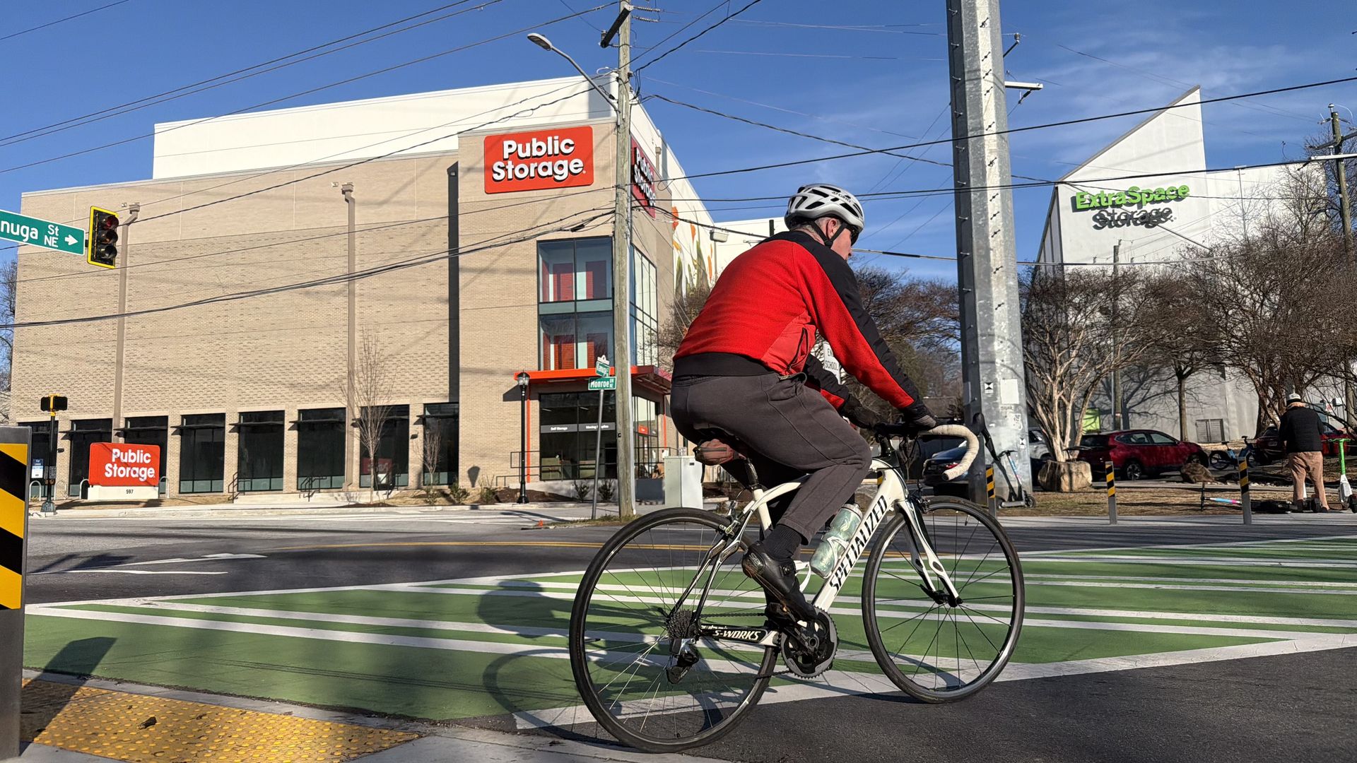 Cyclist in red and black jacket riding a white Specialized bike across a green bike lane intersection near Public Storage and Extra Space Storage buildings under a clear blue sky.