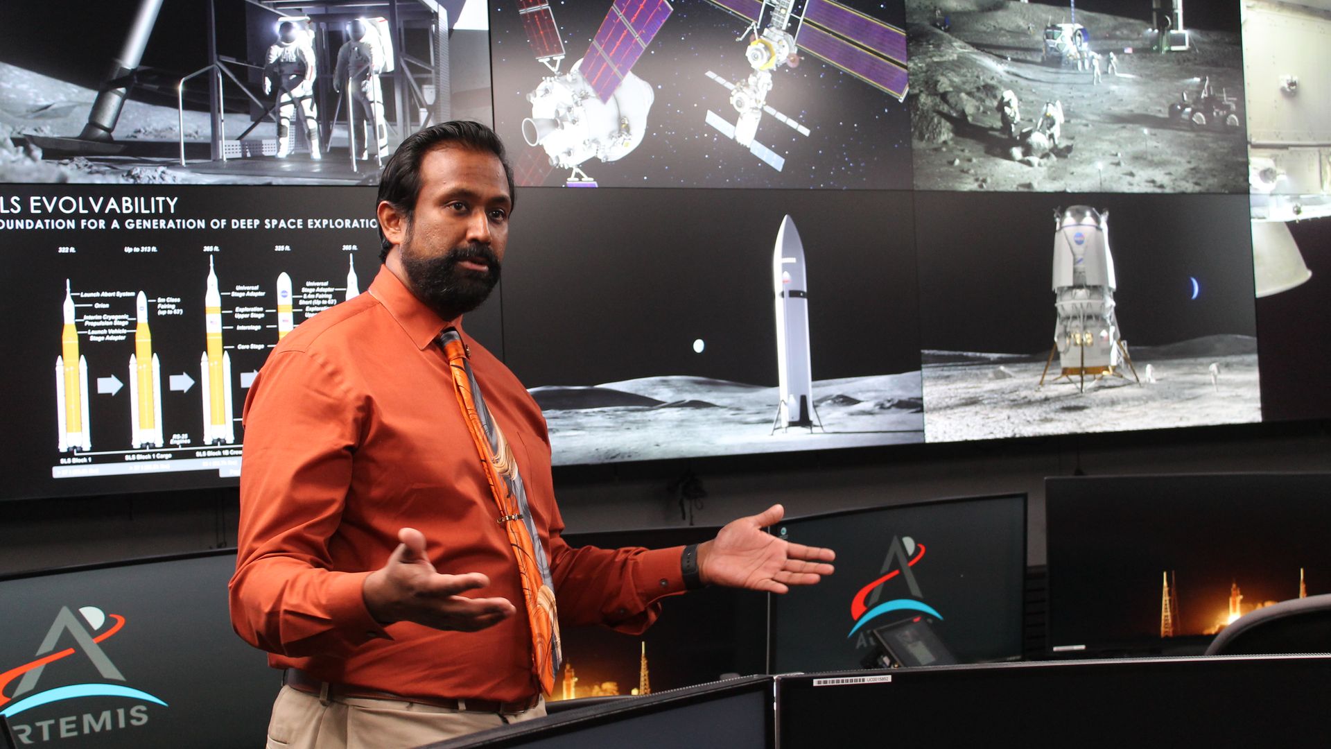 Man in orange shirt and orange space-themed tie giving presentation in front of screens showing lunar and space exploration images, including astronauts, rockets, and satellites, labeled Artemis.