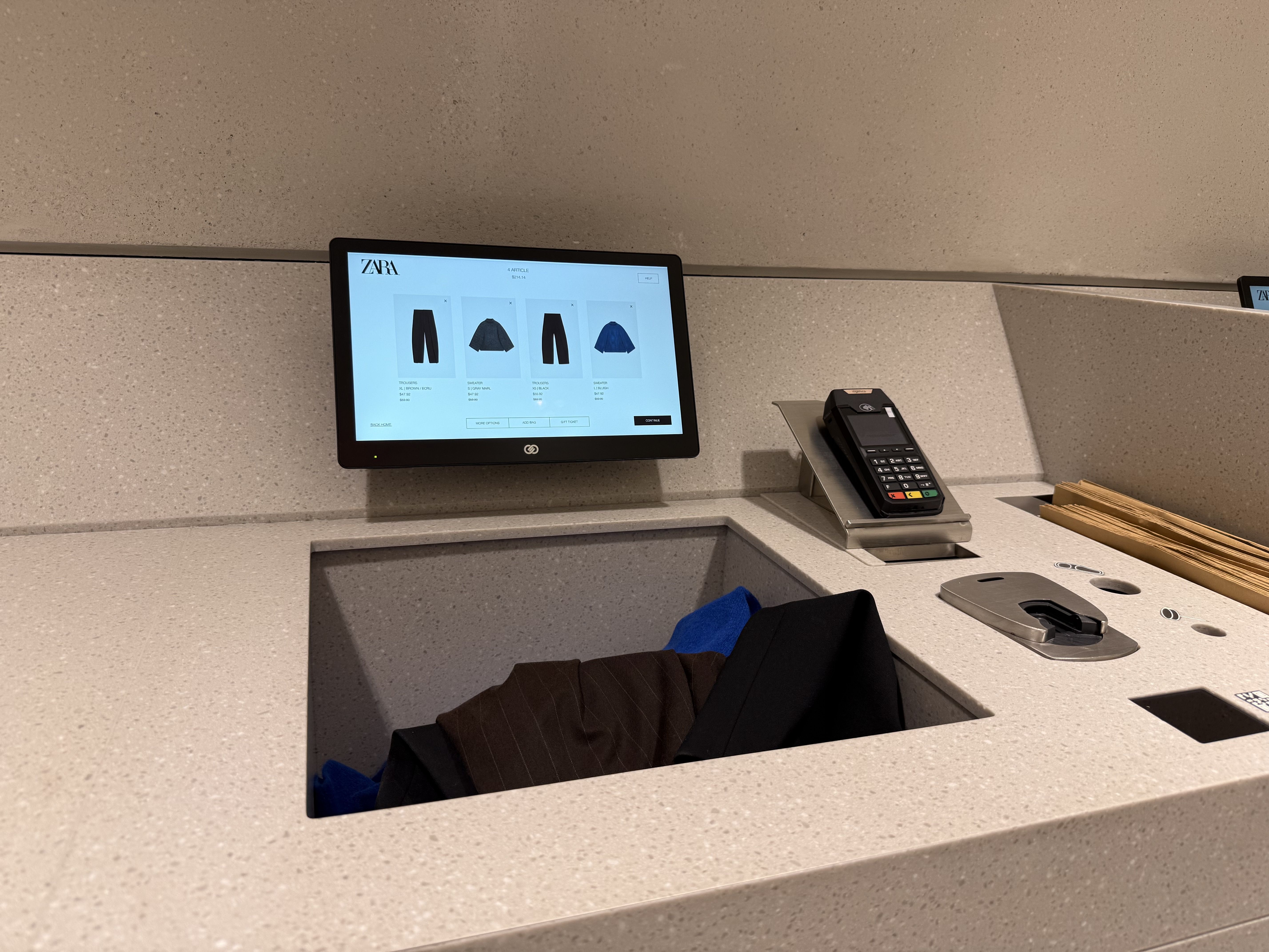 Retail checkout counter with a screen displaying Zara clothing items, a card payment terminal, and a recessed compartment holding folded clothes in brown, black, and blue.