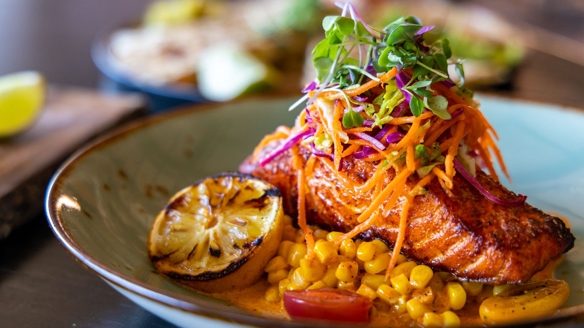 A dinner plate with grilled salmon topped with a fresh salad with a bed of corn and tomatoes underneath.