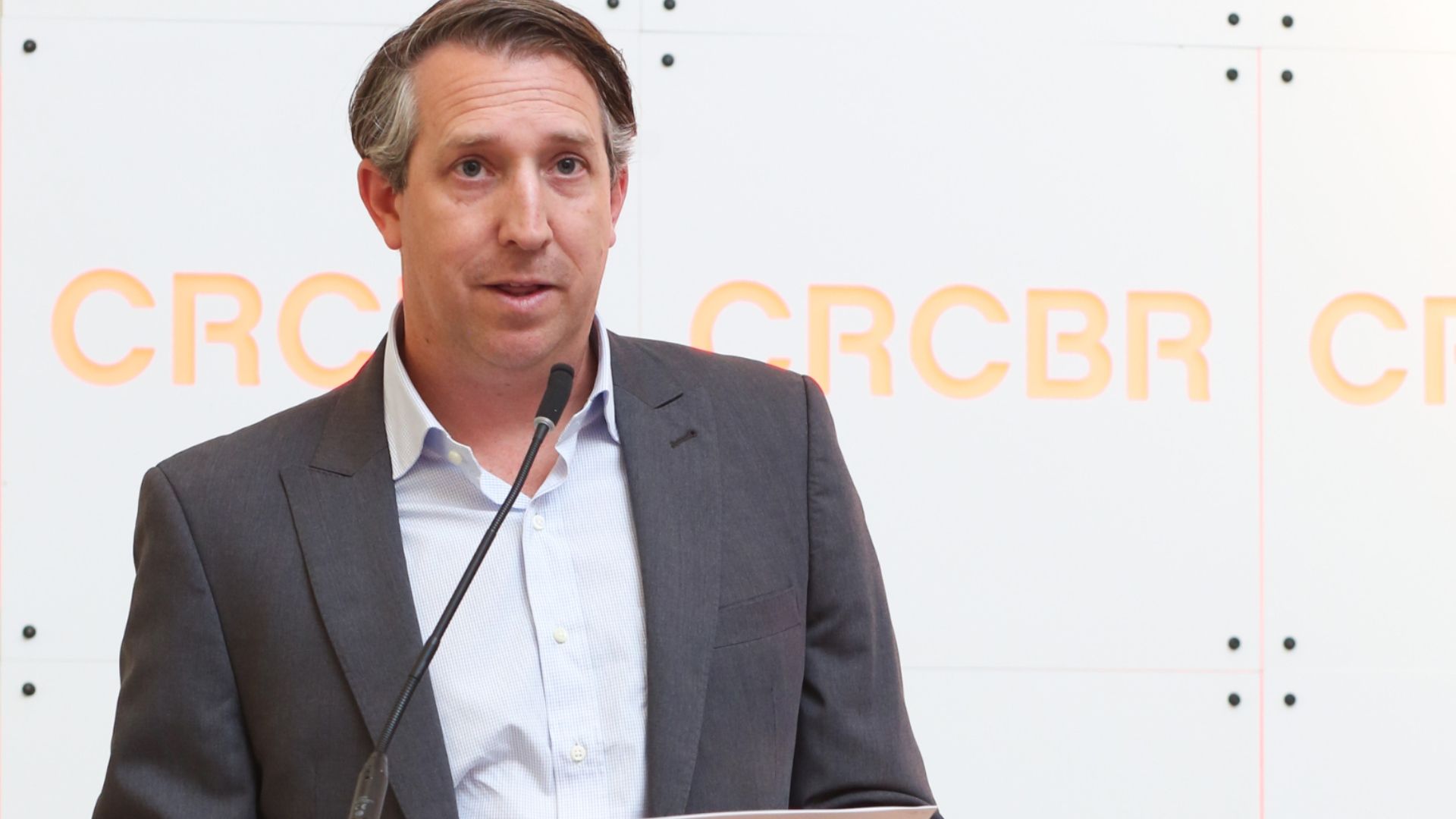 A man in a dark gray suit and light blue shirt stands at a microphone, speaking at a podium, in front of a white wall with large orange letters in the background.