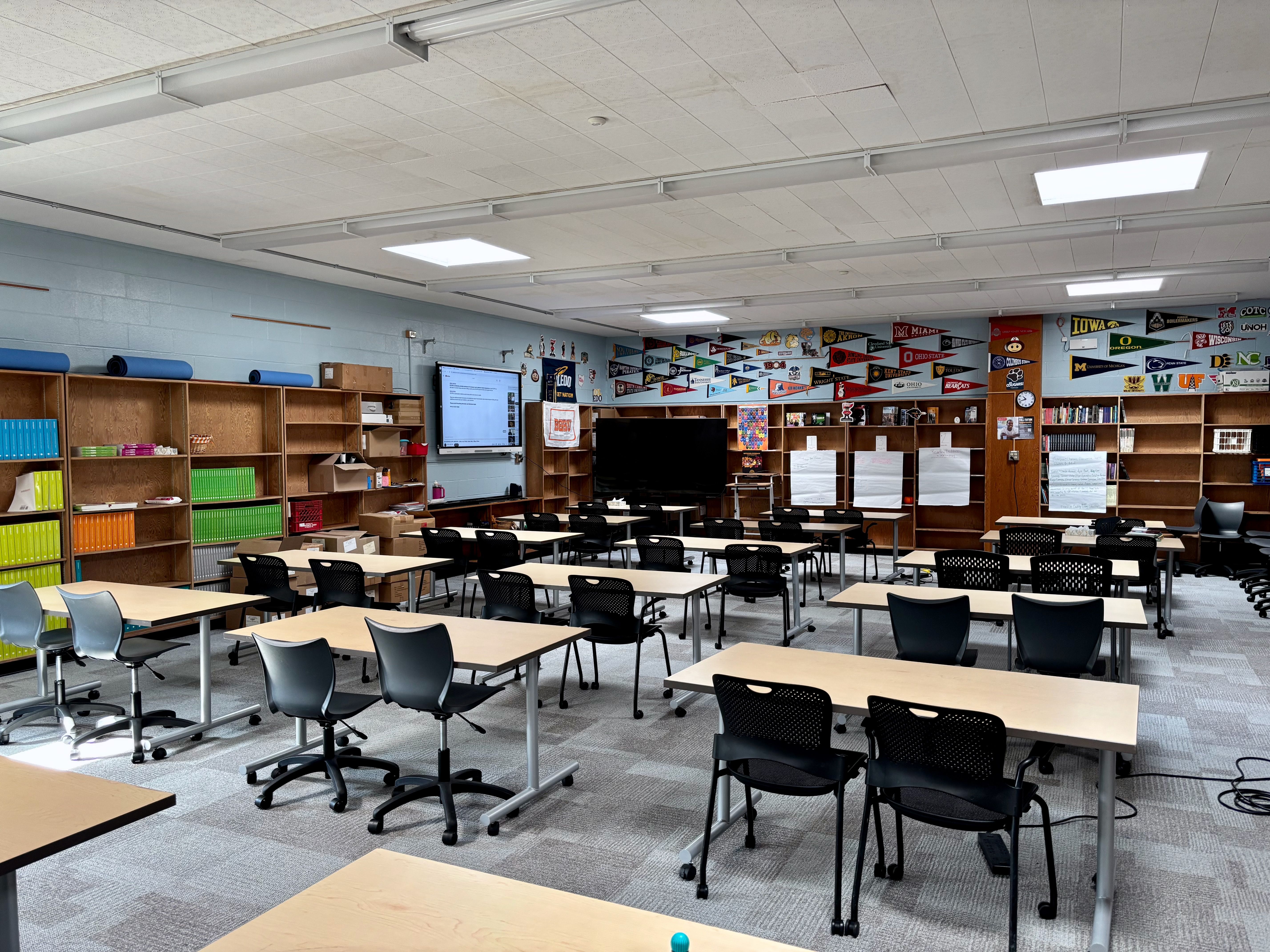 Bright classroom with multiple light wood desks and black chairs arranged in rows. Left wall has tall shelves with colorful books; back wall features pennants, posters, and a large screen.