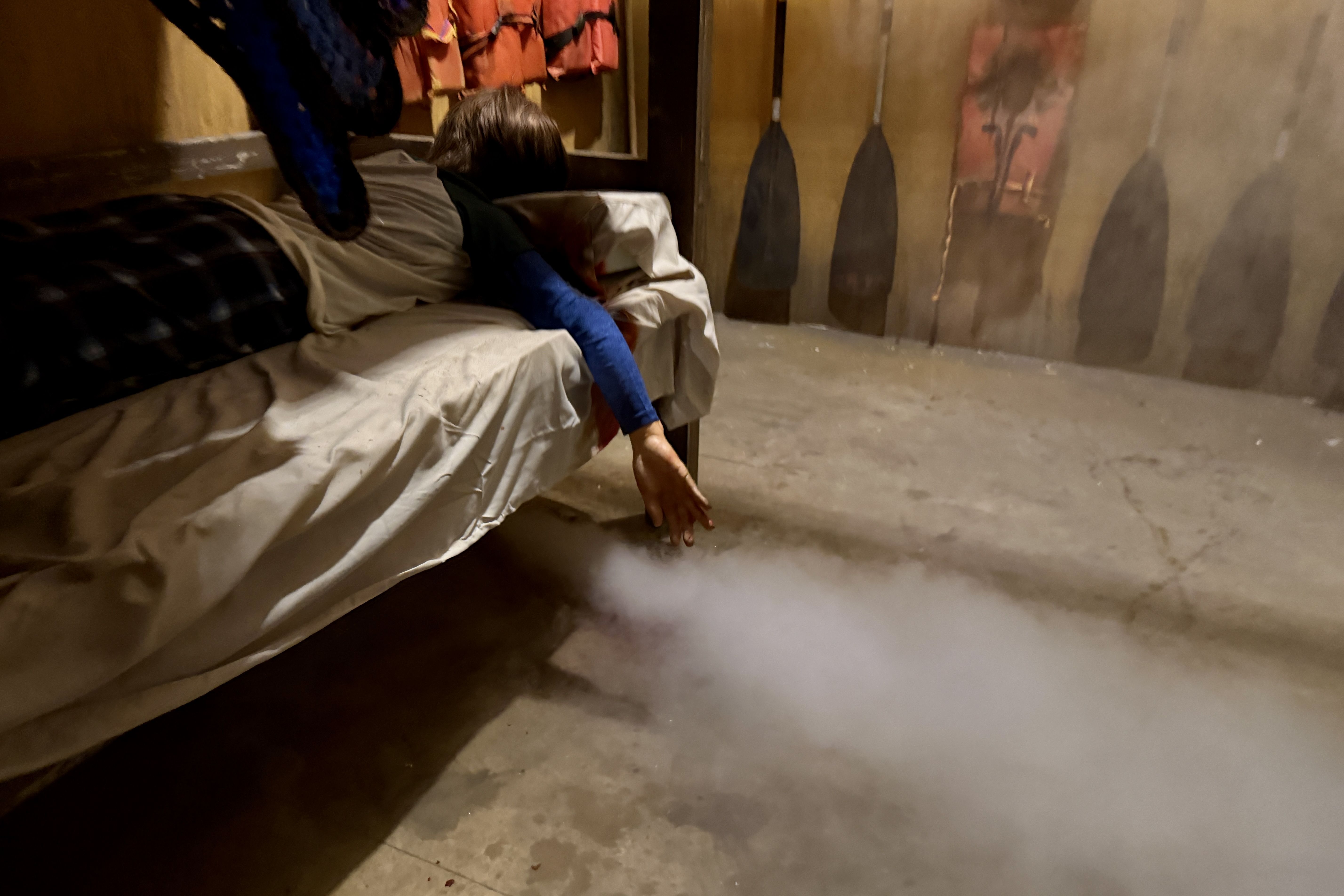 Dim room with a person lying on a bed covered in white sheets; a blue sleeve hangs over the edge as fog swirls on the floor. Jackets hang on the wall beside faded art.