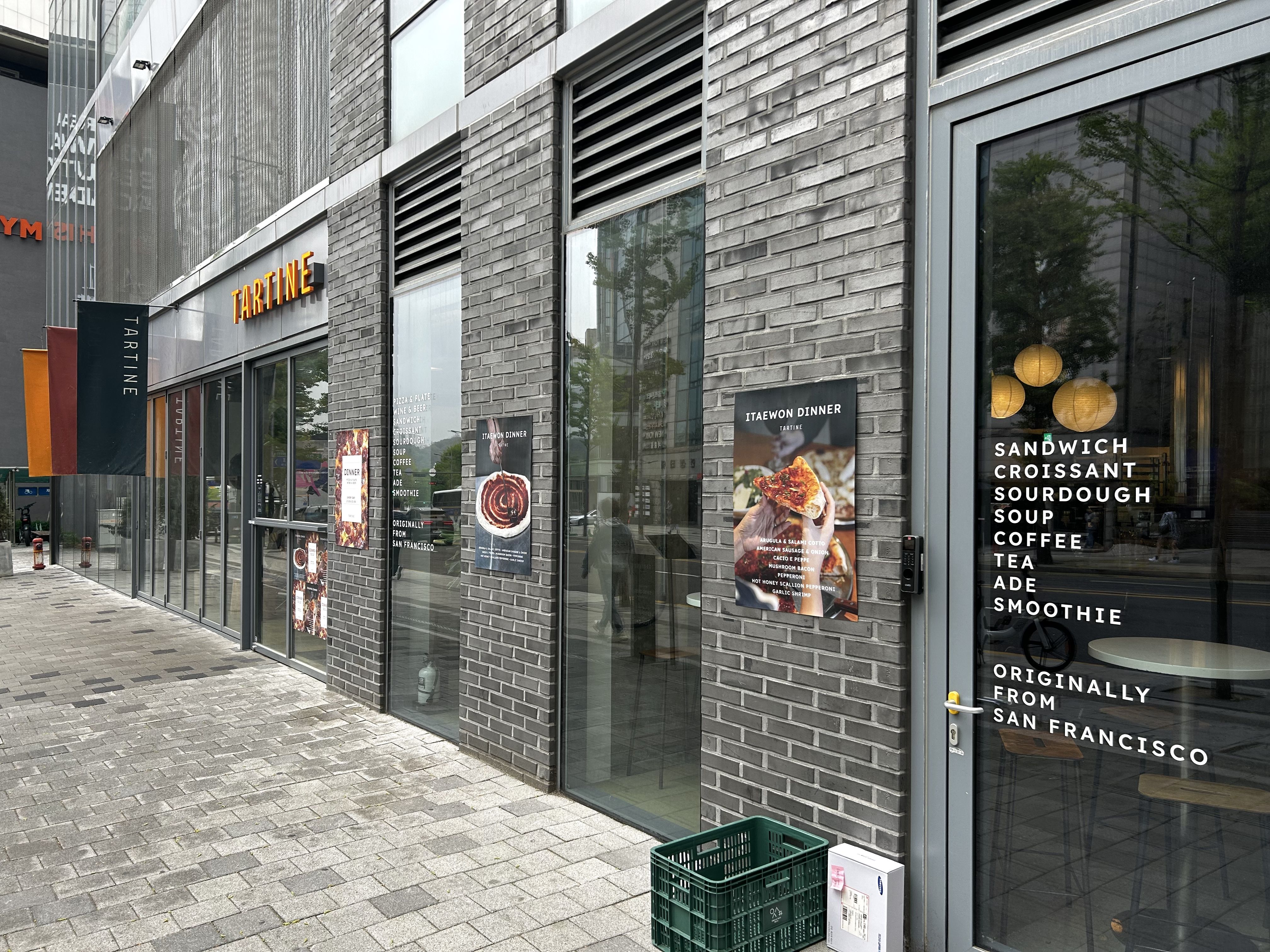 Gray brick storefronts with large glass windows; an orange 'TARTINE' sign above the door; posters of food in the windows; a green crate on the sidewalk; tree reflections visible in the glass.