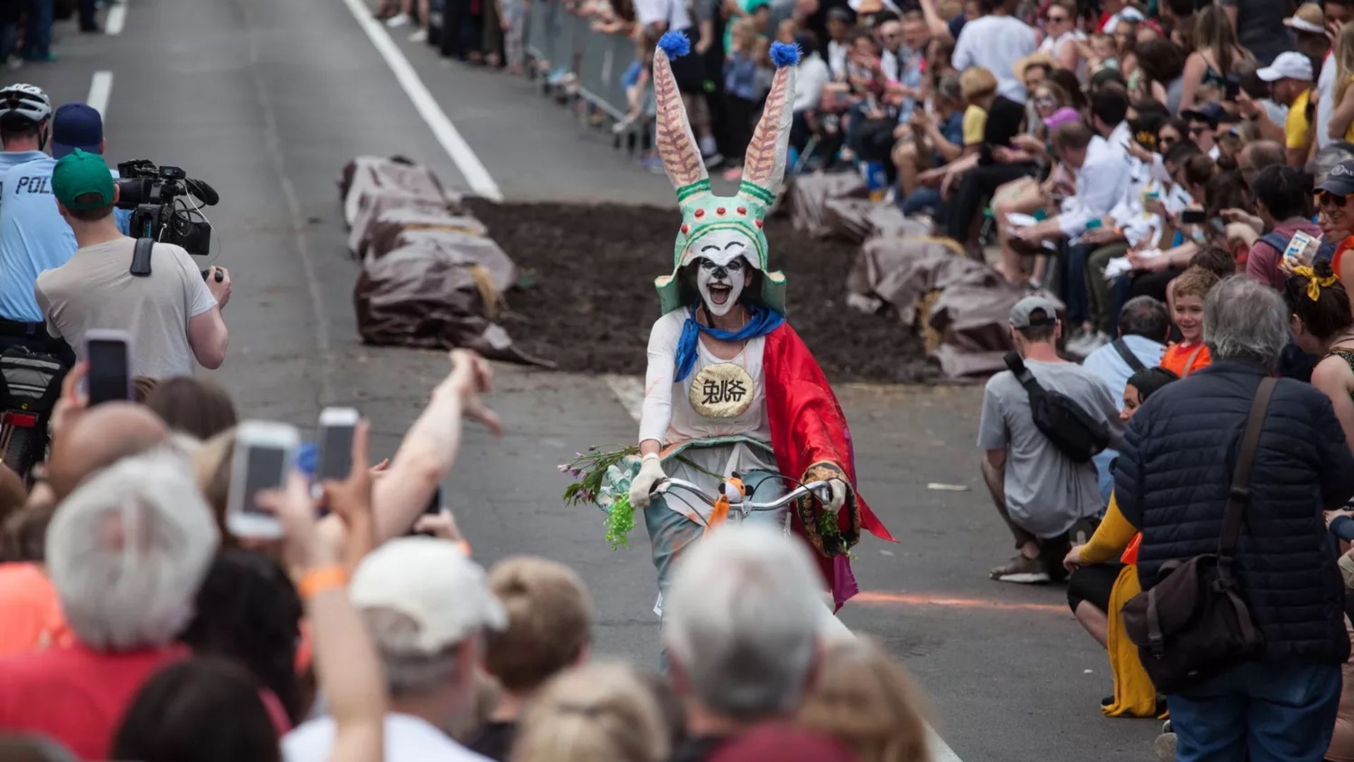 A crowd cheers on a clown riding a bicycle in the street