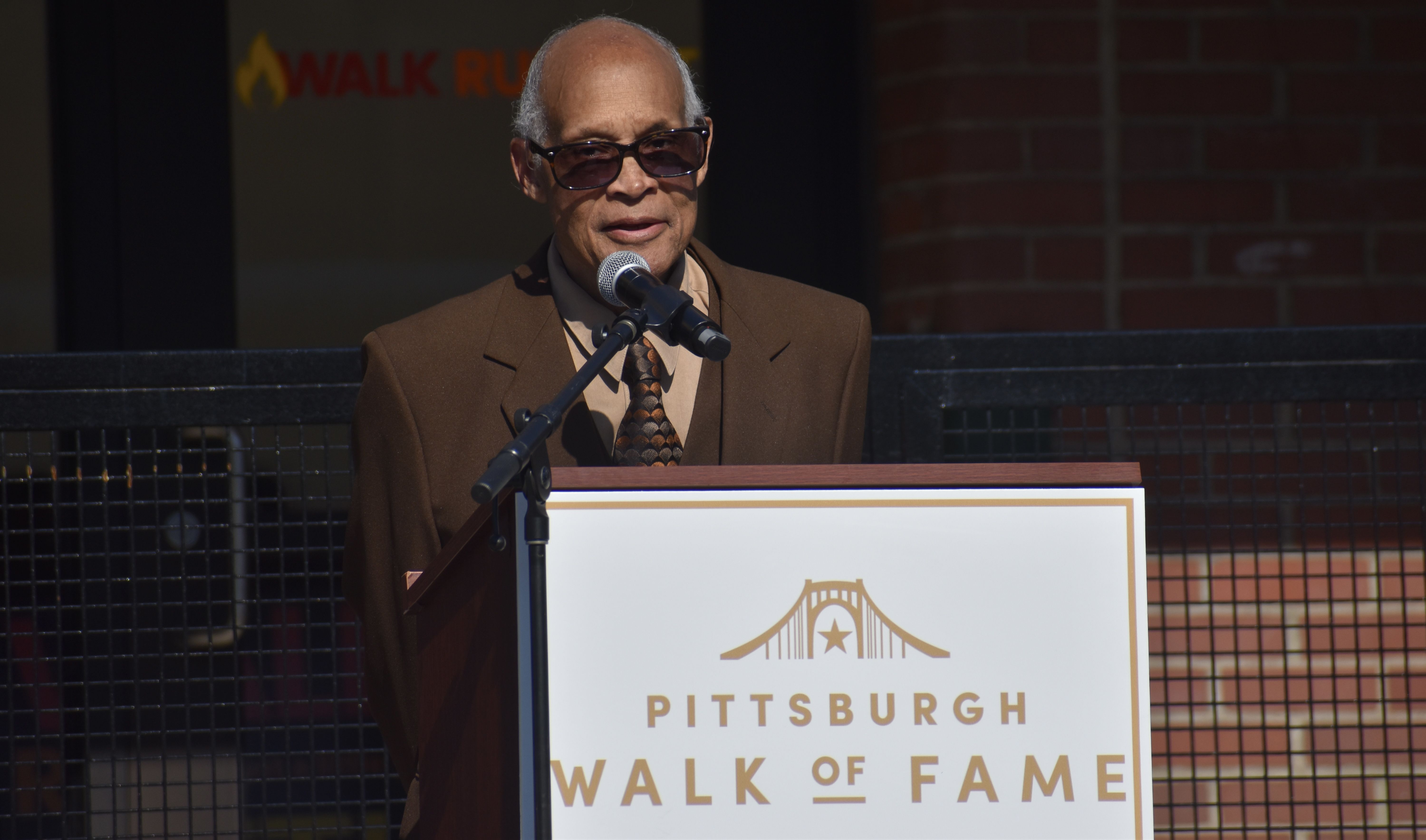 A man in a brown suit and patterned tie wearing dark sunglasses speaks into a microphone at a podium. A "Pittsburgh Walk of Fame" sign with a gold bridge emblem is visible in the background.