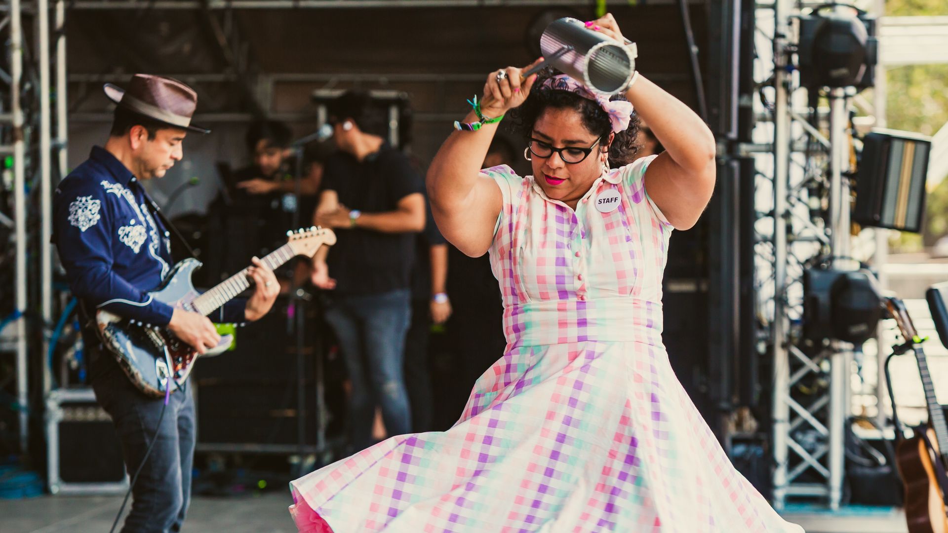 Stage scene with a staff member in a pink plaid dress dancing energetically, wearing glasses and a badge. A guitarist in a blue shirt and hat plays in the background amid stage equipment.