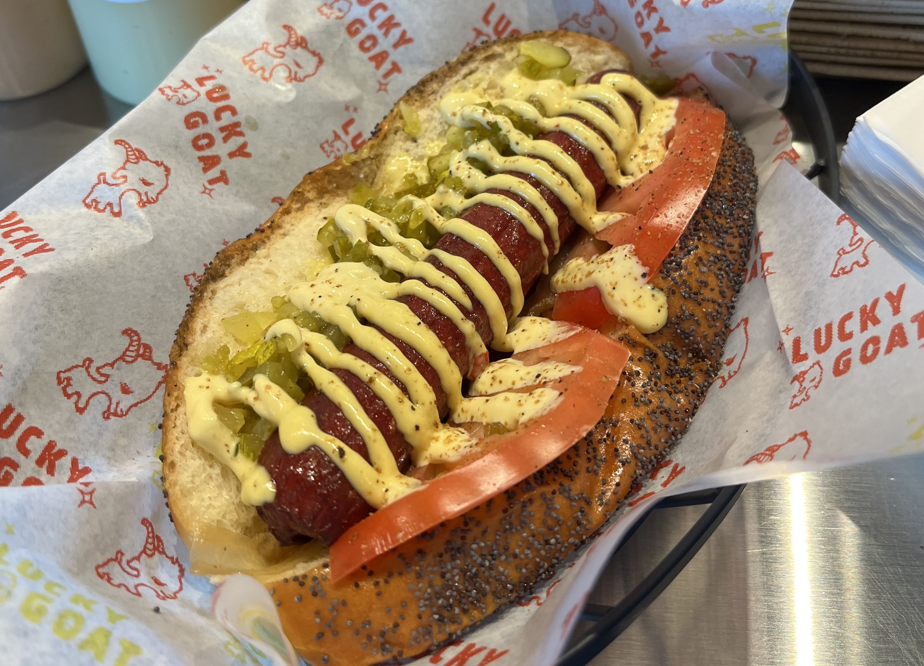 Hot dog in a poppy seed bun with mustard, relish, tomato slices, and onions on Lucky Goat branded paper.
