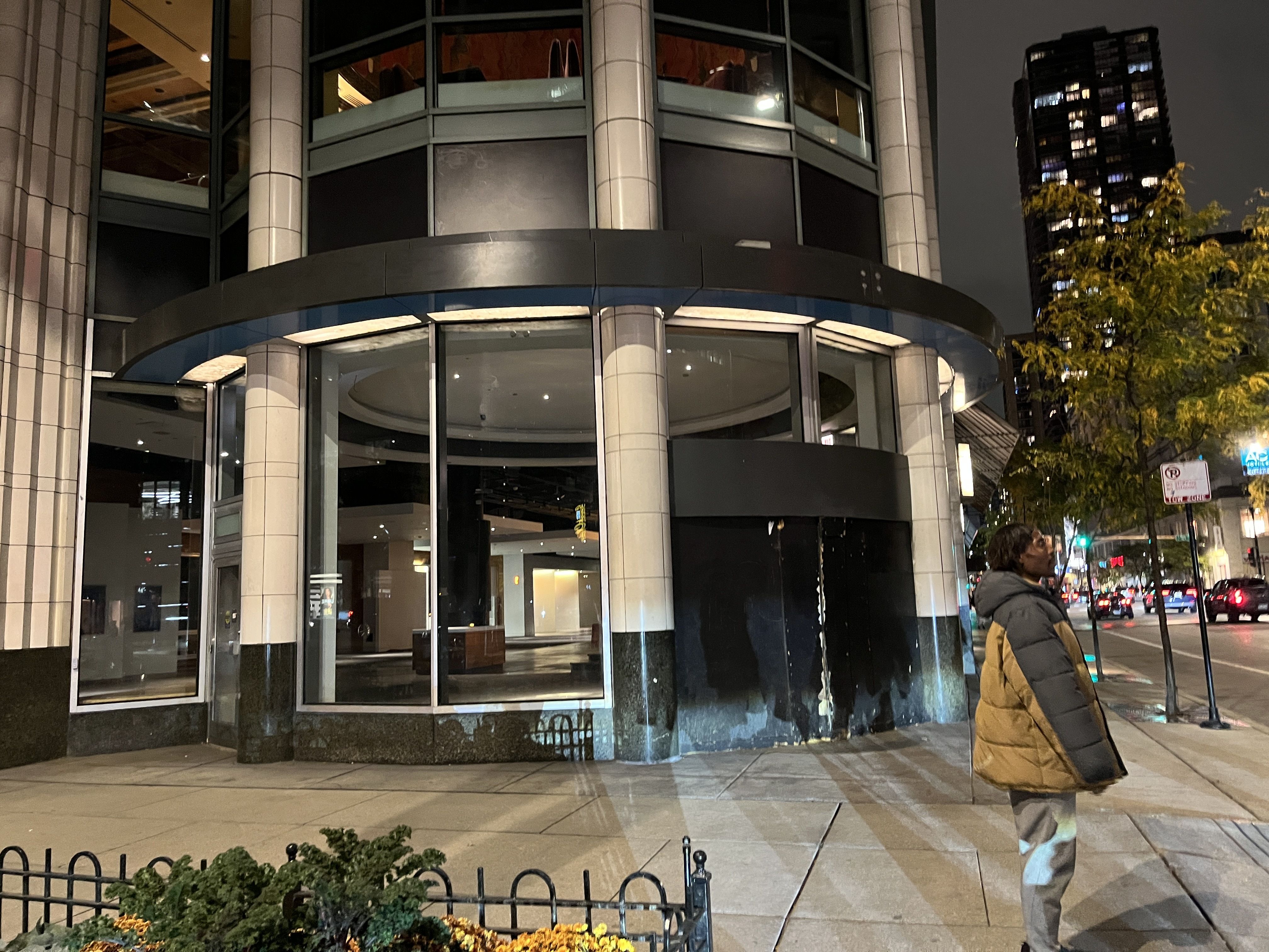 Nighttime street view of a modern round building with large glass windows and beige pillars. A person in a puffy brown and black jacket stands near the corner, with city lights and trees in background.