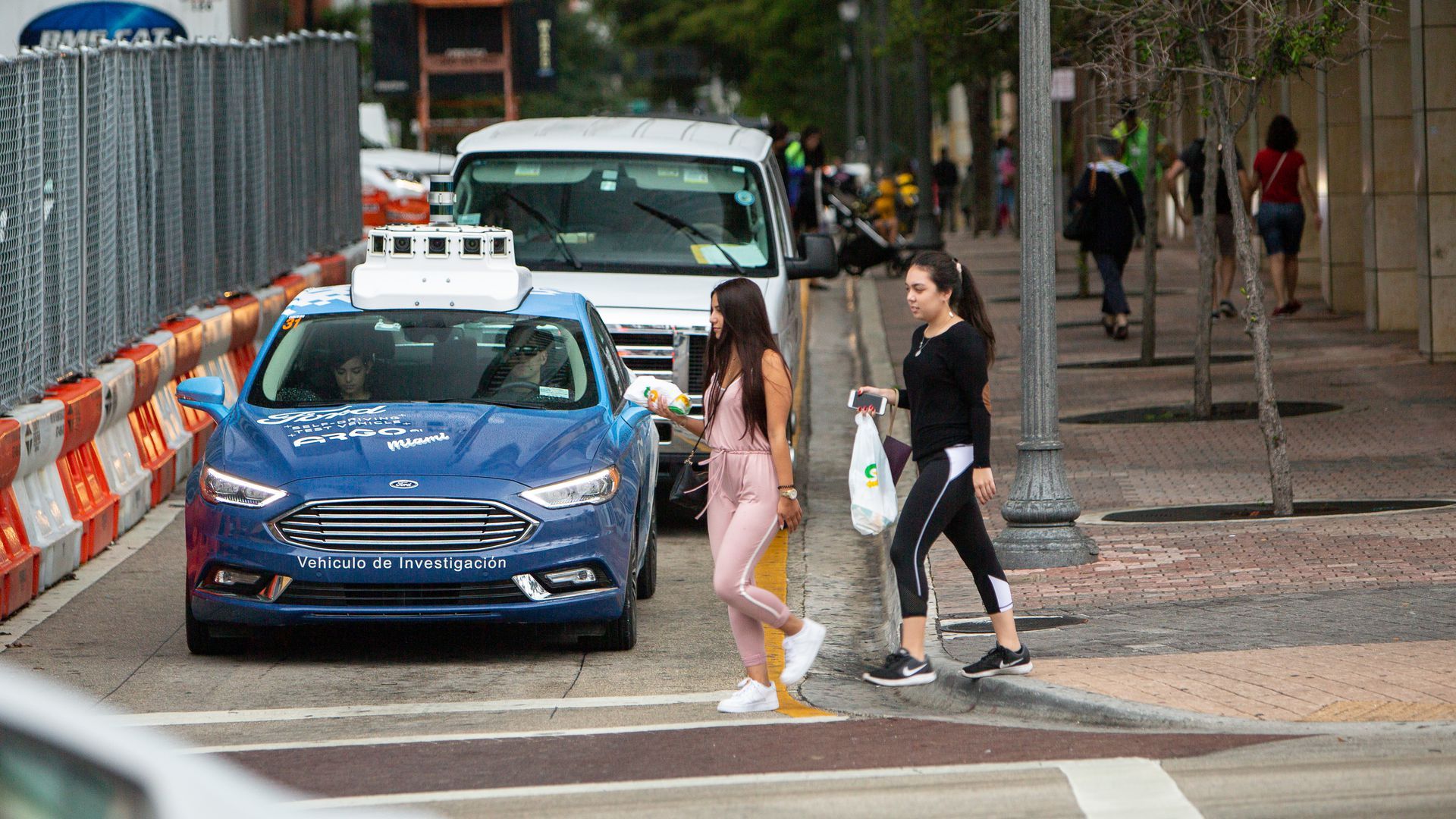 In this image, pedestrians cross a city street in front of a blue sedan. 