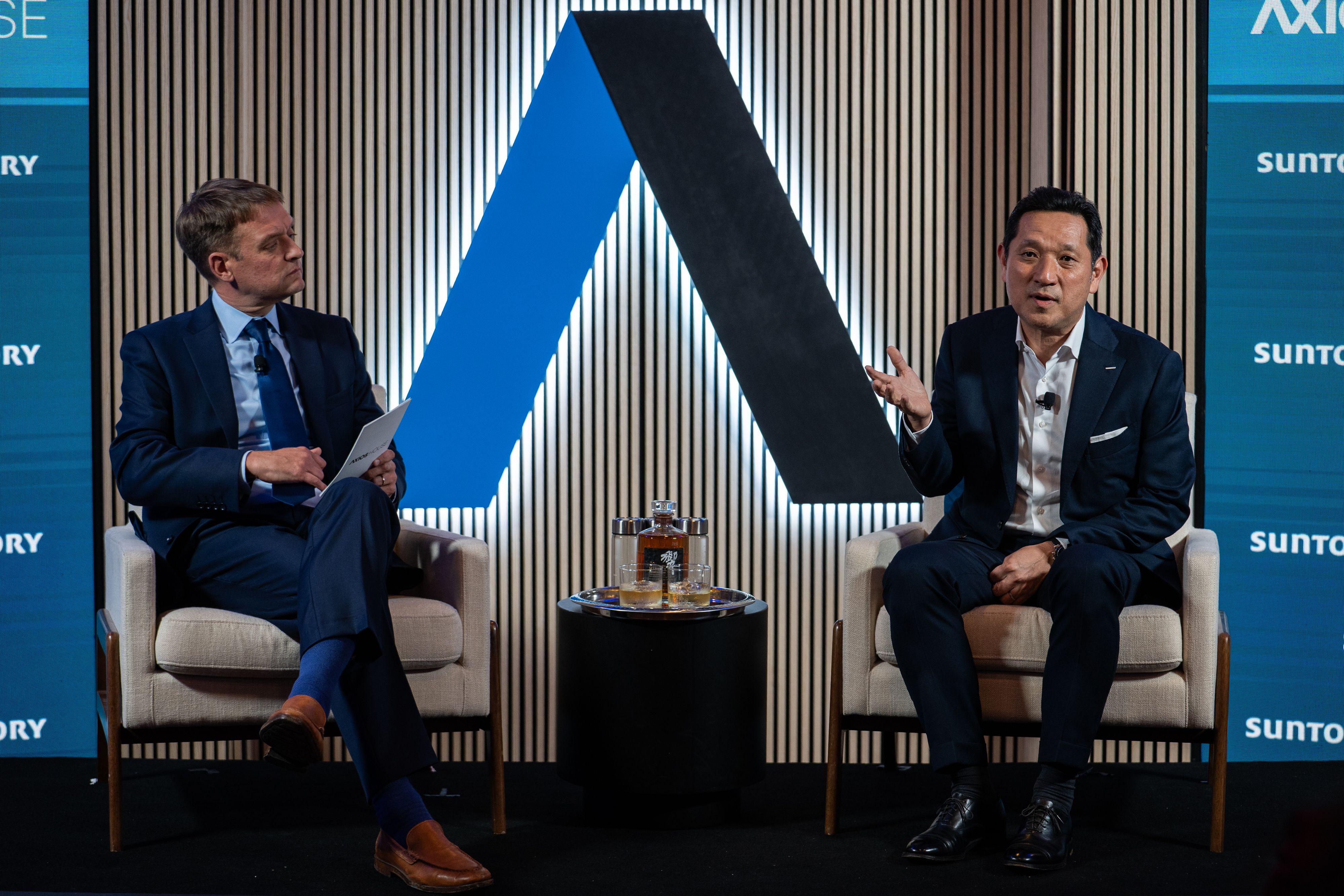 Two men in dark suits sit on stage in cream chairs, facing each other. A large blue and black Axios logo sits between them; drinks on a small table; blue panels display Suntory logos.