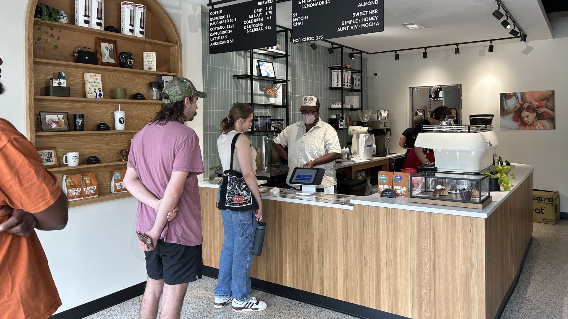 Two people wait in line at a modern and bright coffee shop with a black-and-white simple menu over the counter and shelves lined with cameras and mugs
