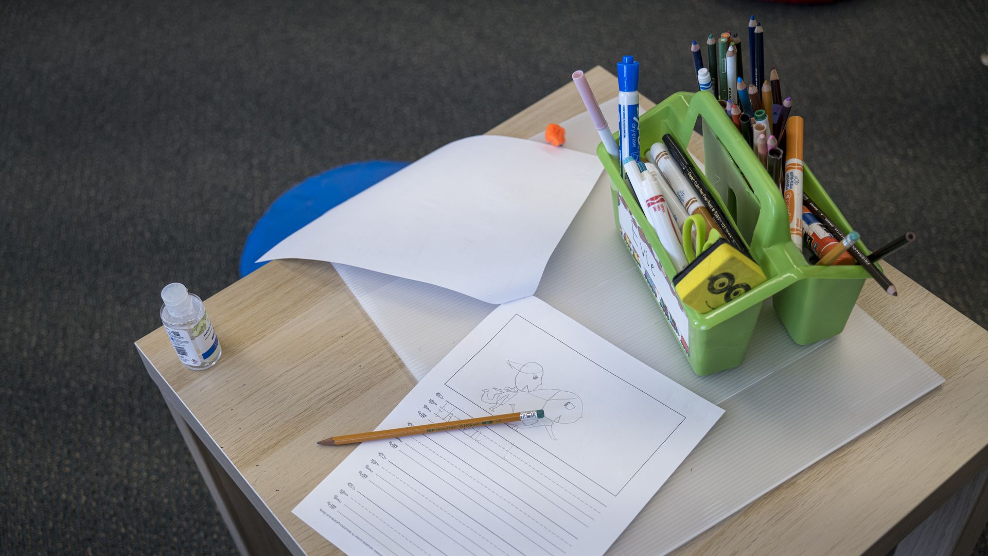 Photo of a classroom desk with a packet of papers and a tub of markers 
