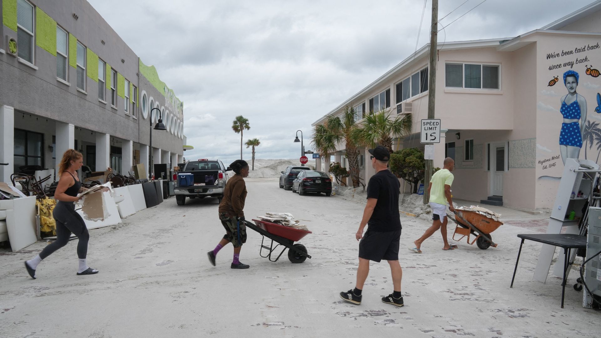 Two people with wheelbarrows and one holding a hunk of drywall cross a road covered in sand.