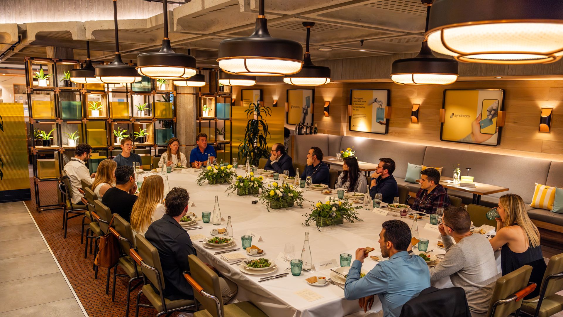 Group of people seated around a large rectangular dinner table with white tablecloth, green plants centerpiece, green glasses, and plates with food in a warmly lit restaurant with modern decor.